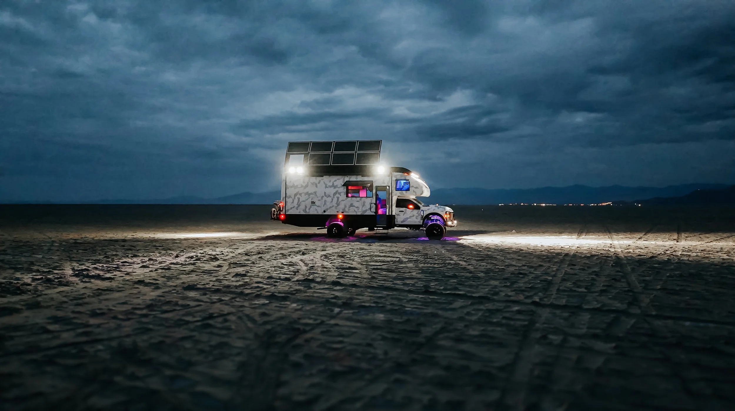 A converted van with solar panels on the roof parked on a sandy beach at dusk, with a cloudy sky and distant city lights on the horizon.