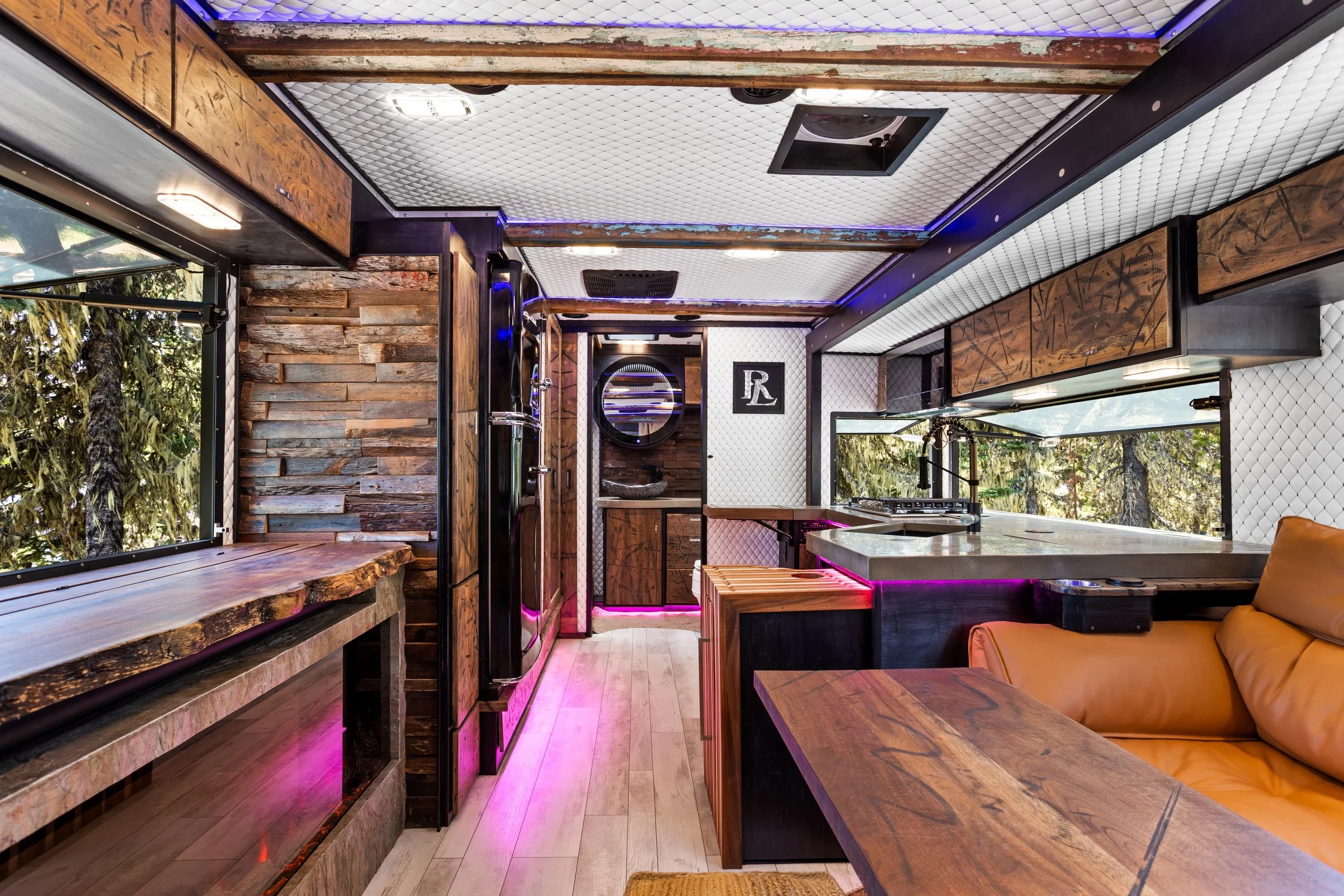 Interior of a modern rustic kitchen with wooden and stone elements, large windows showing trees outside, and ambient purple lighting.