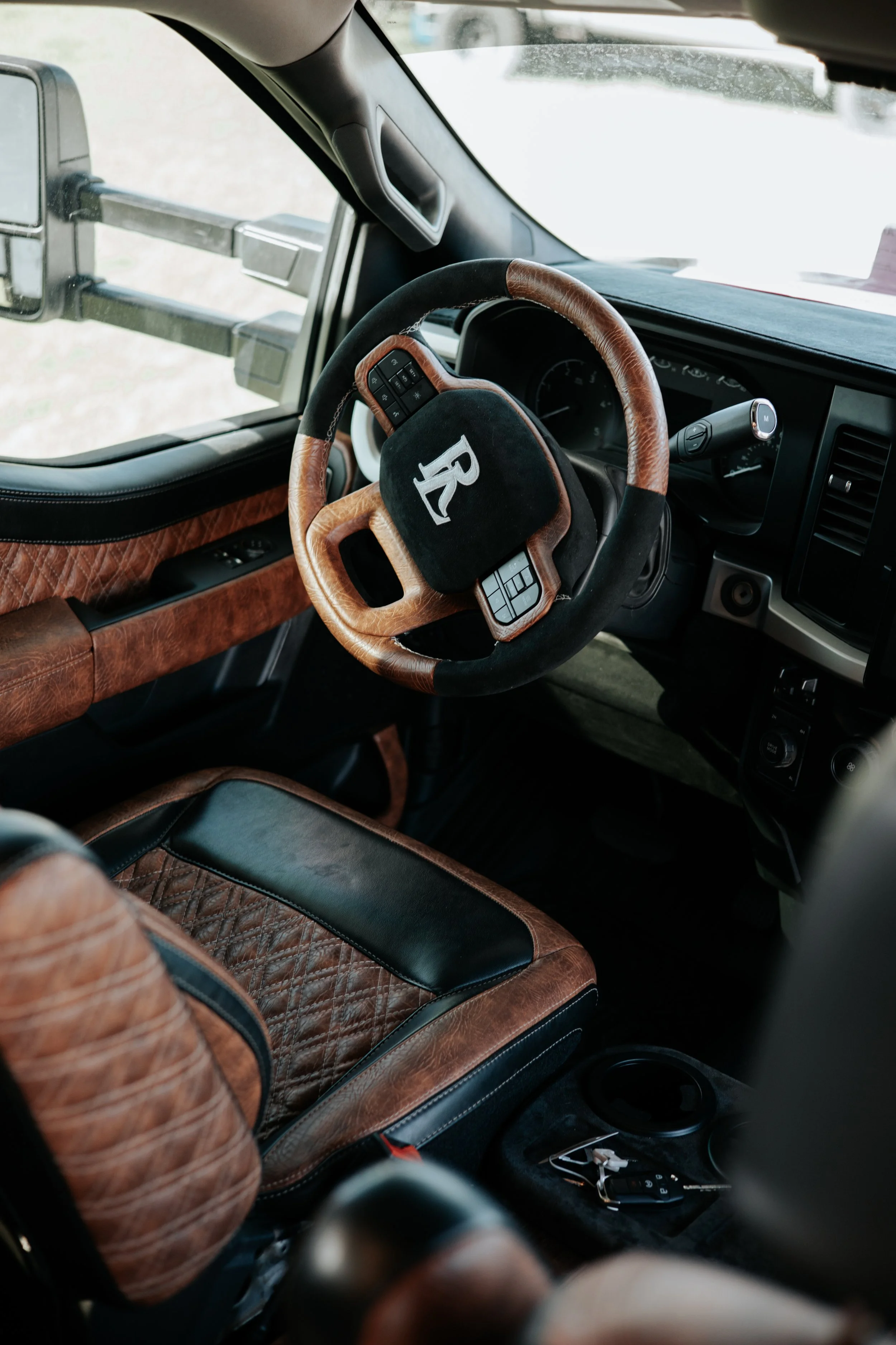 The image shows the interior of a vehicle with a brown leather and black fabric quilted steering wheel cover, a matching quilted door panel, and a black dashboard. The driver’s seat has a matching quilted brown leather and black fabric design. On the floor, there are various items, including a car key, a black electronic device, and some other small items.