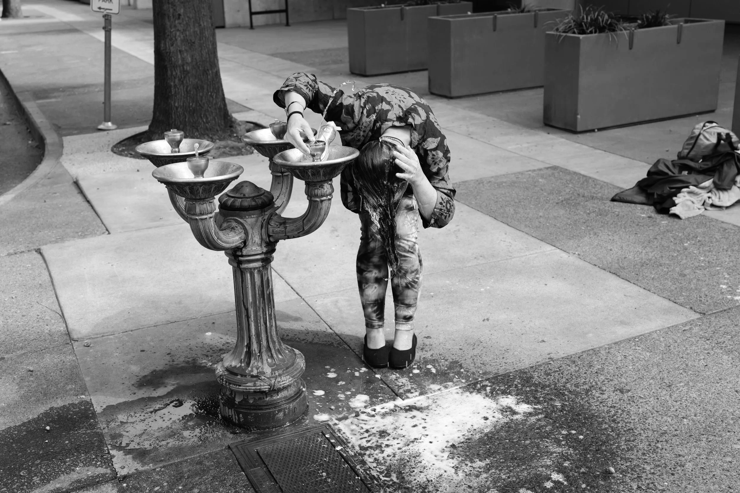 A person with long hair wearing camouflage jacket and tie-dye pants, bending over, washing their hair at a water fountain on a city sidewalk with a tree, sidewalk, planters, and clothes on the ground nearby.