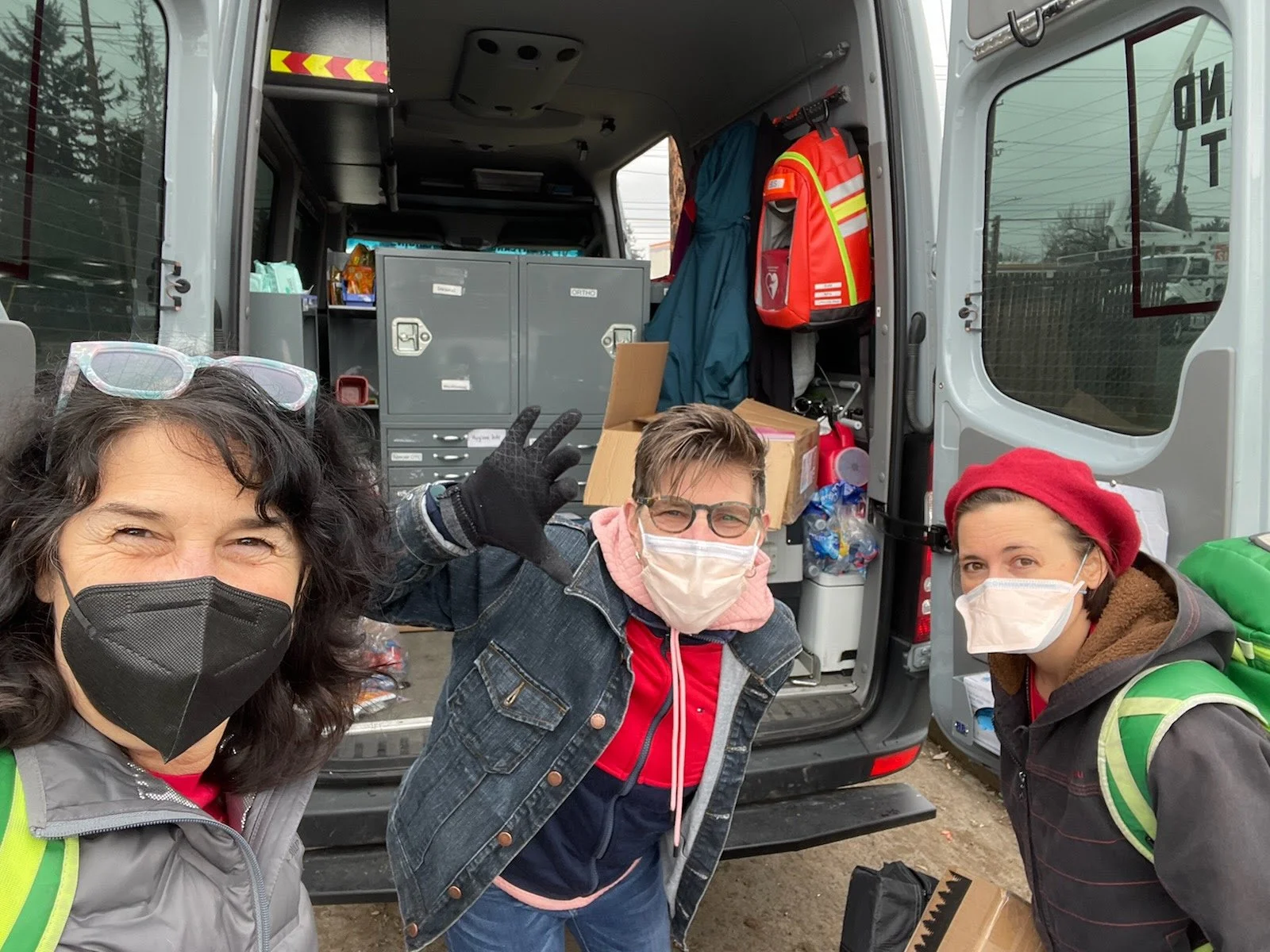 Three people wearing face masks and safety vests smiling in front of an emergency response van filled with supplies, with trees and power lines in the background.