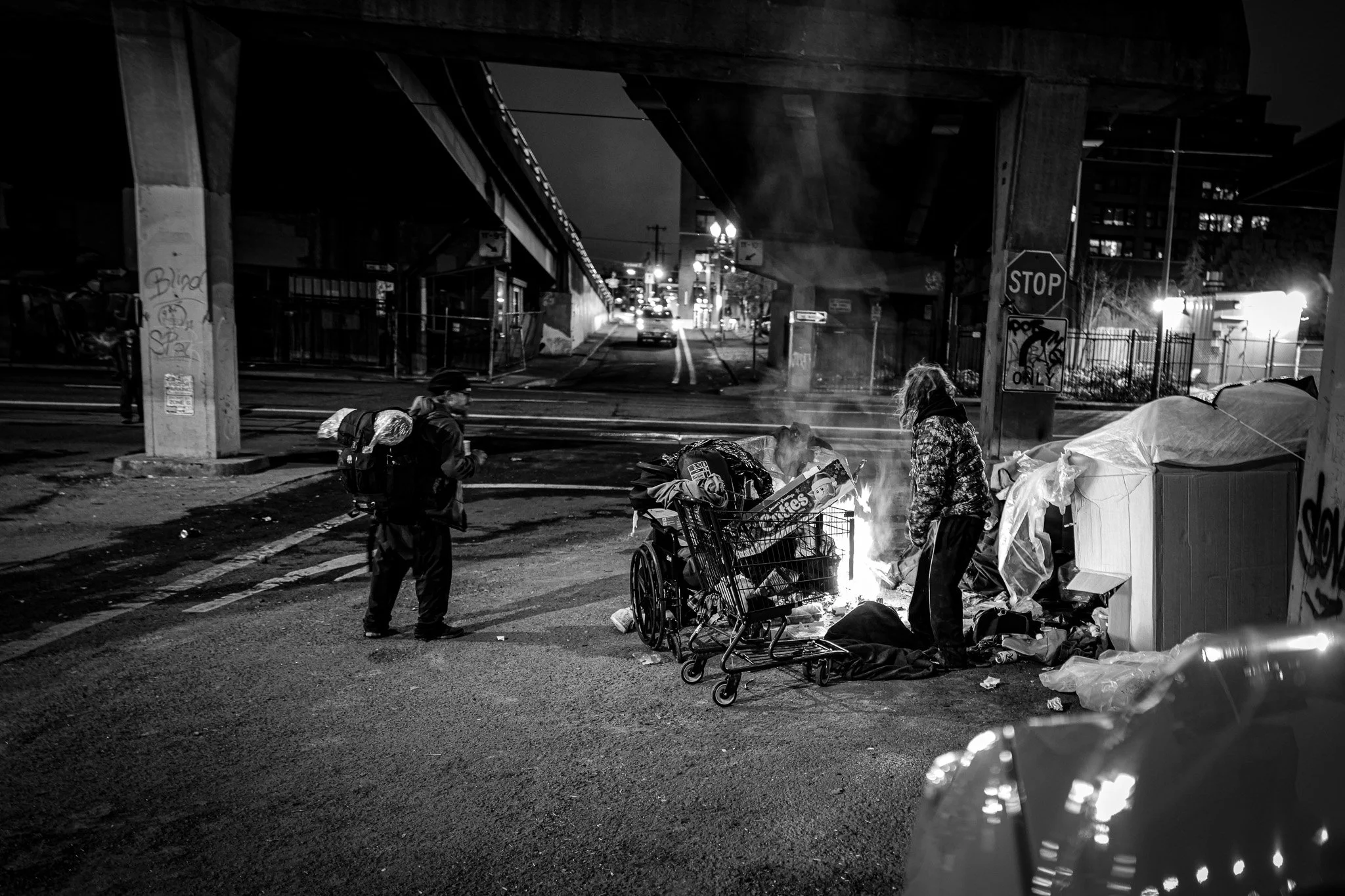 Nighttime urban scene with two people standing near a small fire among trash and shopping carts under a bridge, with city lights and streets in the background.