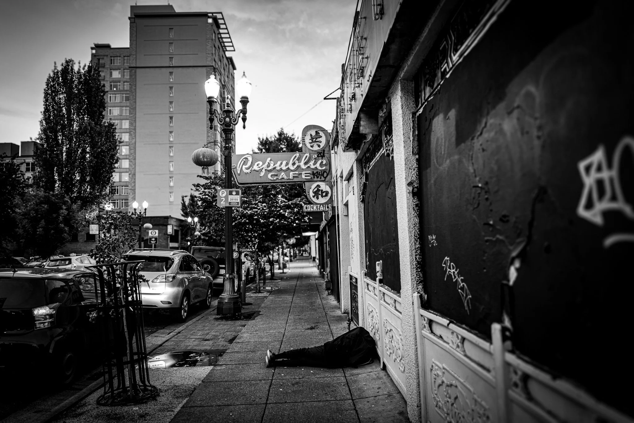 Black and white street scene with parked cars, a sidewalk, a storefront with graffiti on the right, and illuminated street lamps. A high-rise building and trees are in the background.