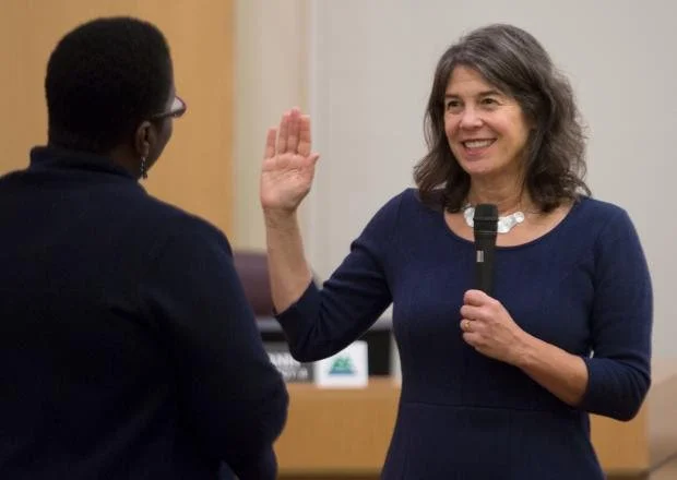 A woman (Sharon Meieran) raising her right hand and smiling, holding a microphone in her left hand, engaging in a swearing-in ceremony with another woman.
