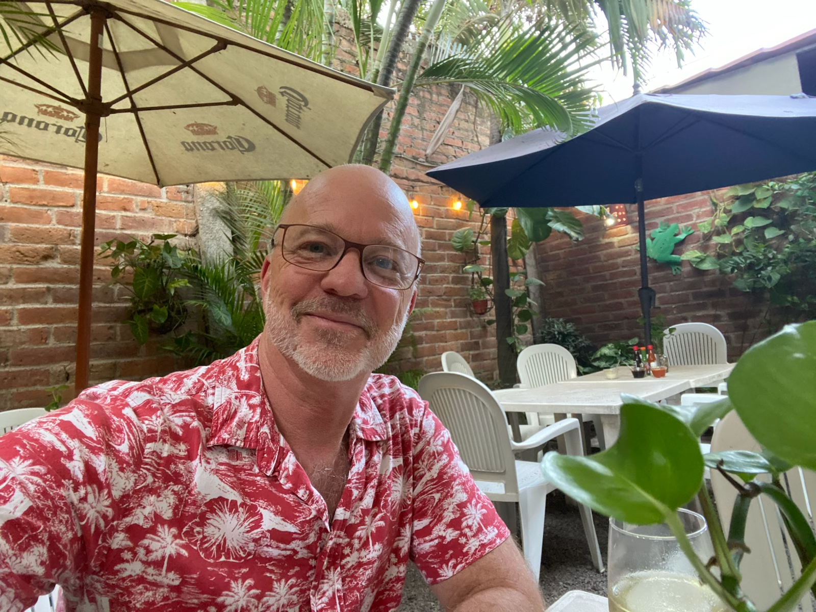 A man with glasses, white hair, and a beard taking a selfie at an outdoor patio. He is wearing a red Hawaiian shirt with white floral patterns. The background has brick walls, green plants, and white tables and chairs with umbrellas, one beige with a Corona logo and the other dark blue.