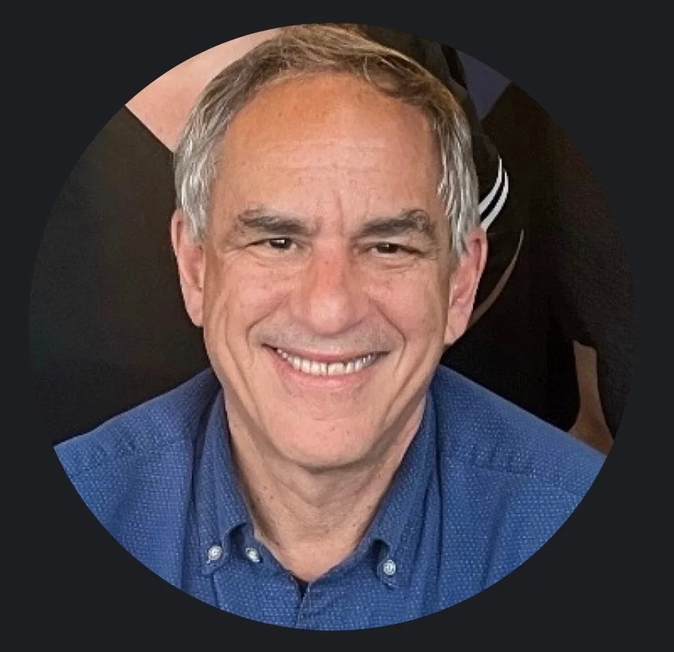 Close-up of a smiling middle-aged man with gray hair, wearing a blue collared shirt.