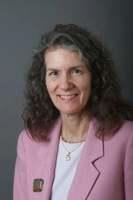 A woman with shoulder-length gray curly hair, smiling, wearing a pink blazer, white top, a gold necklace, and a lapel pin, against a gray background.
