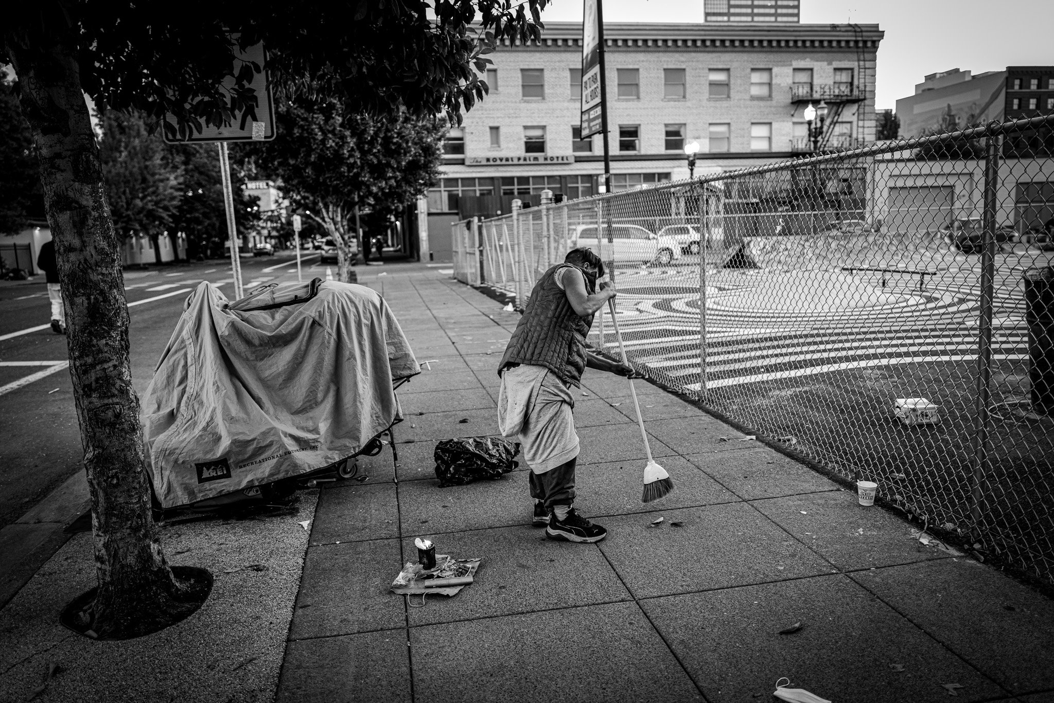 A woman sweeping the sidewalk near a chain-link fence in an urban area at dusk. There are parked cars, trees, and buildings in the background.