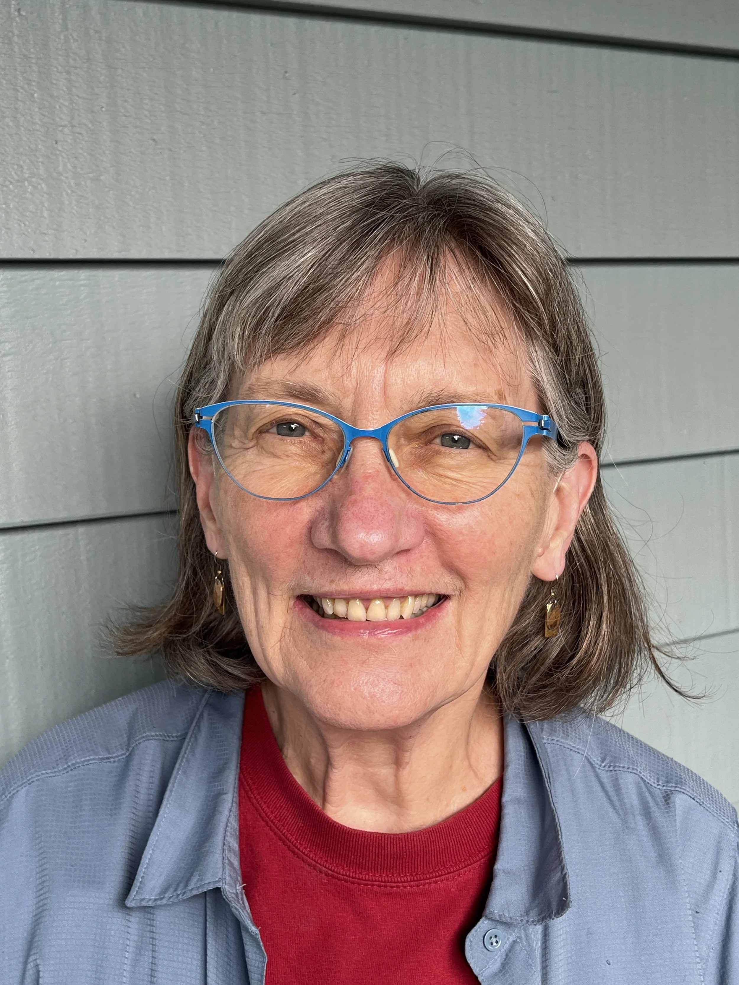 A woman with short gray hair, glasses, and earrings, smiling with teeth showing, dressed in a red shirt and a light blue jacket, standing in front of a gray horizontal panel wall.