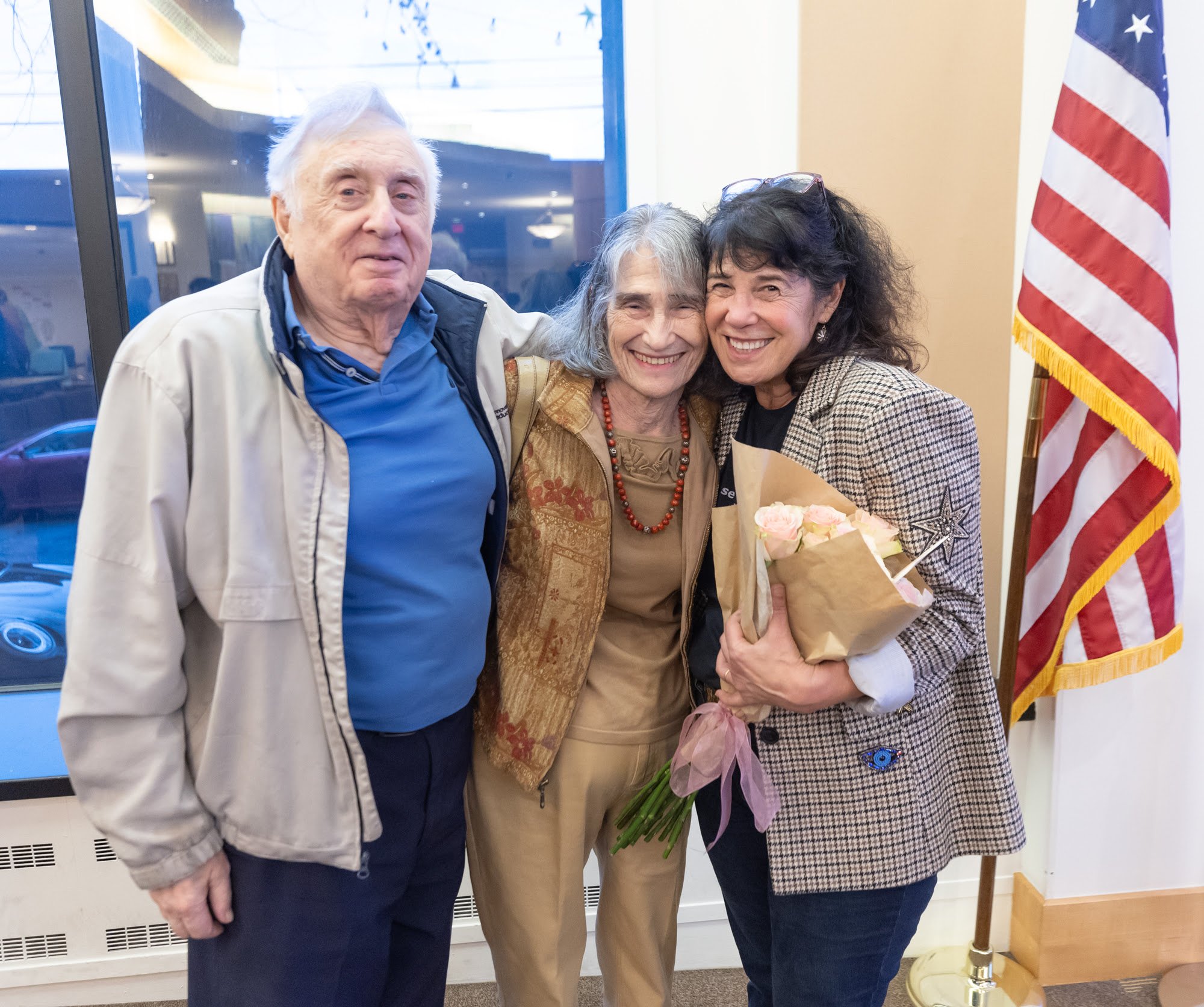 Three people, two women and one man, standing together and smiling, with a woman holding a bouquet of flowers, in front of a window and an American flag.