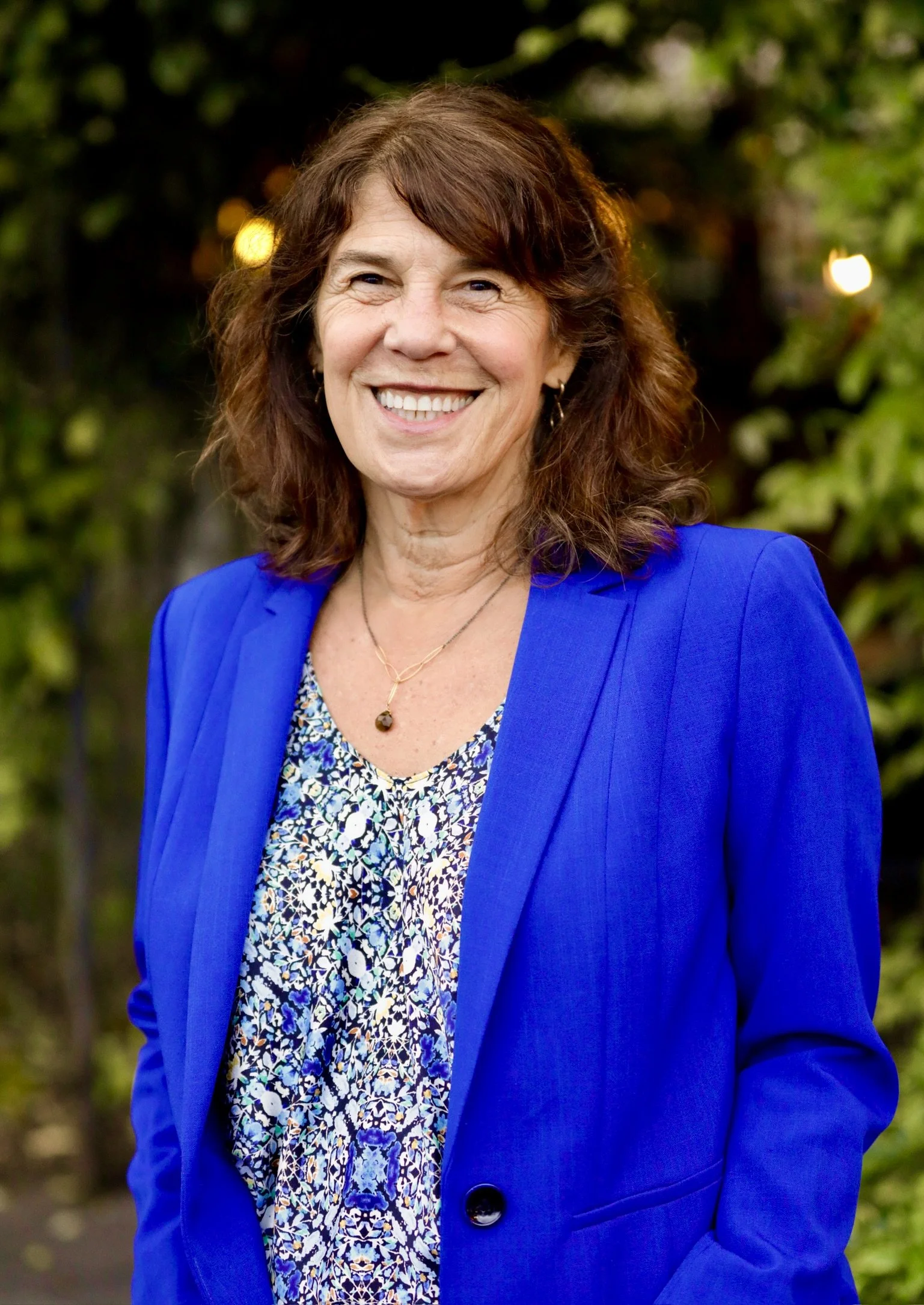 A smiling middle-aged woman (Sharon Meieran) with brown, curly hair wearing a bright blue blazer over a patterned blouse, standing outdoors with blurred greenery in the background.