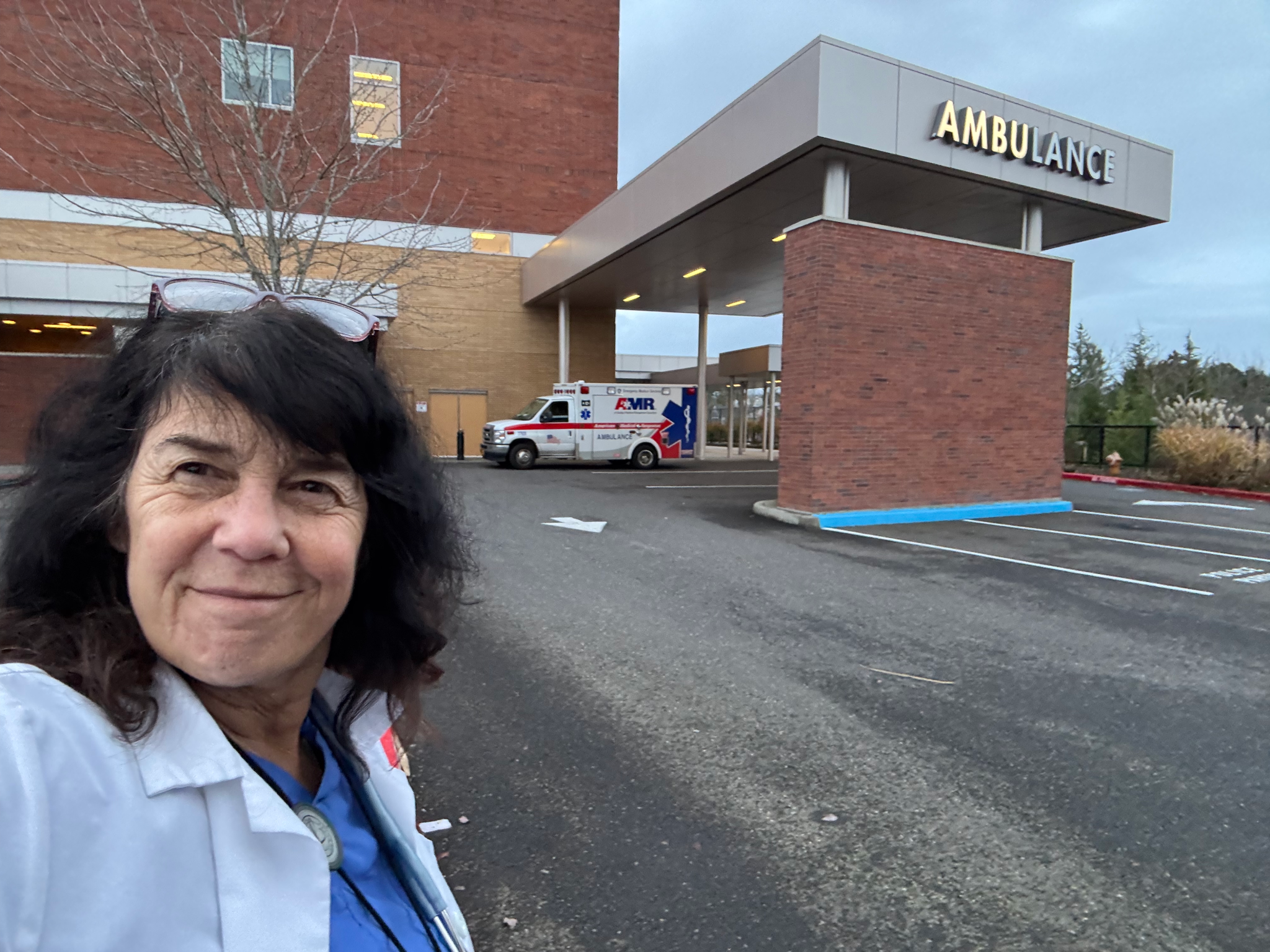 A woman (Sharon Meieran) in a white jacket standing outside a hospital with an ambulance nearby, under a sign that reads 'AMBULANCE' on the building.
