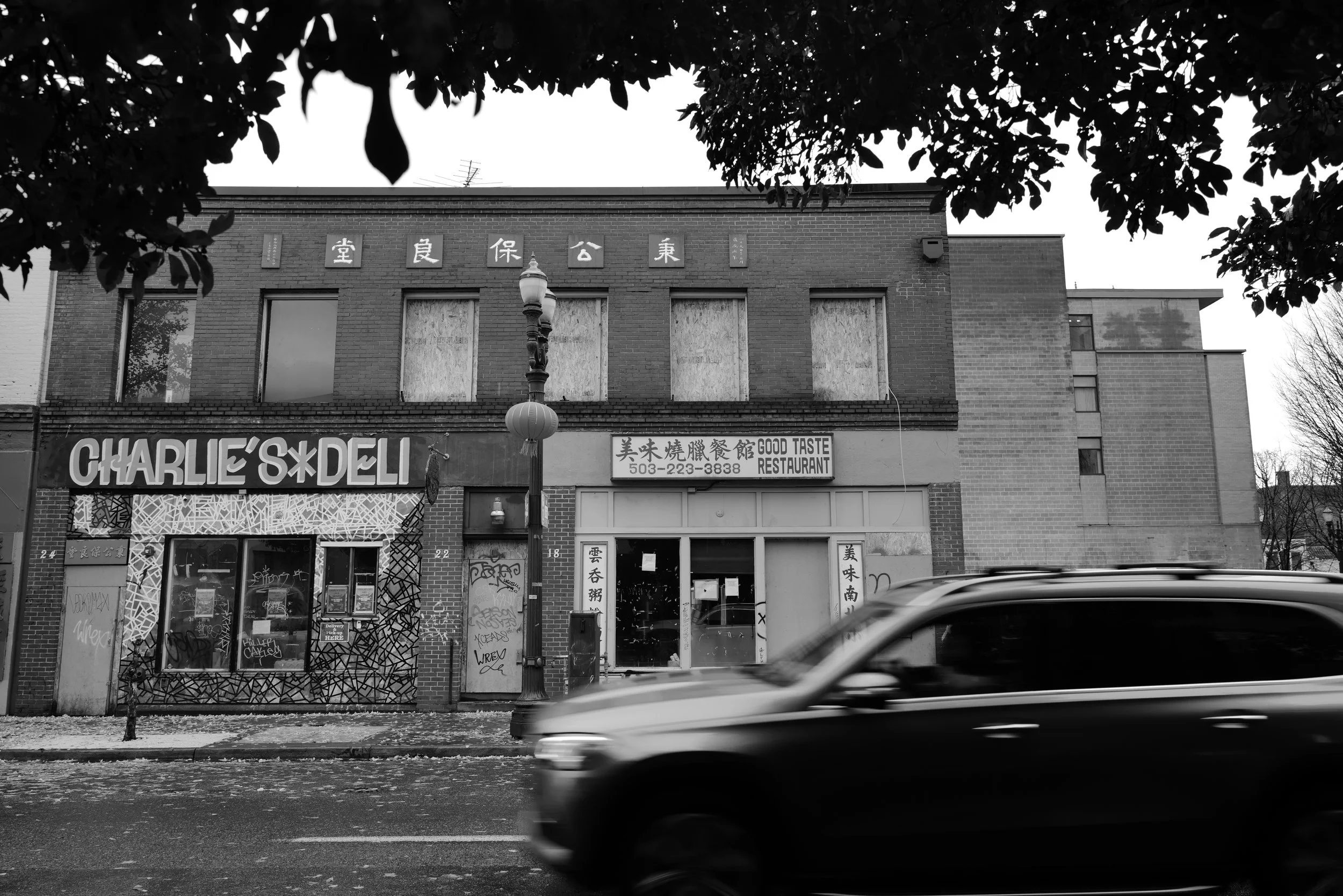 Black and white photo of a brick building with boarded-up windows, graffiti on storefront, and Chinese characters on signs. Moving car blurred in foreground, trees overhead