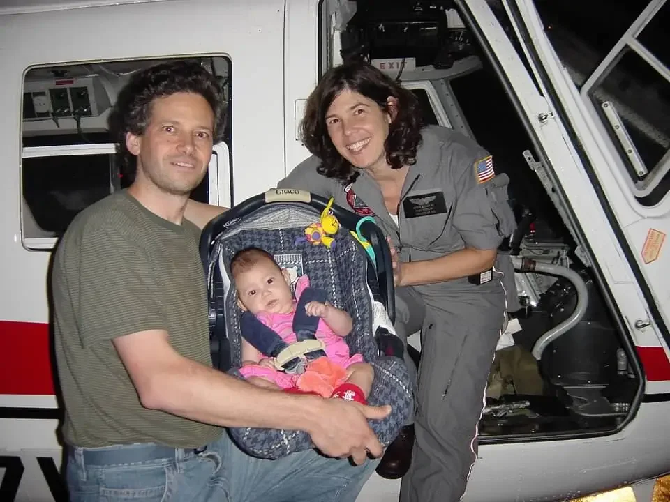 Sharon Meieran & husband, one in a gray uniform and the other in a green shirt, are holding a baby in a car seat in front of a helicopter at night. The woman is smiling widely.