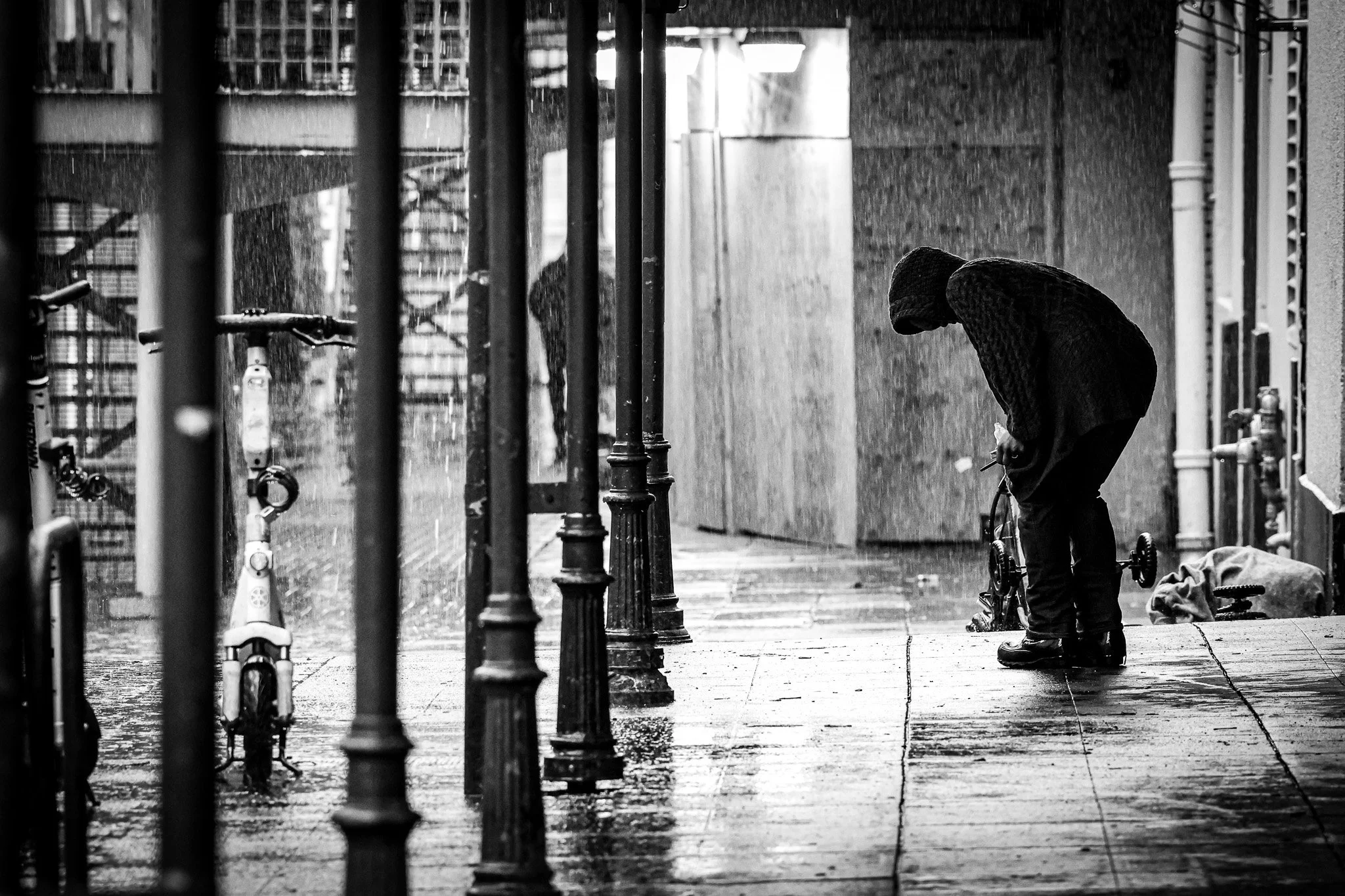 A person wearing a hooded jacket and boots is riding a bicycle on a wet city sidewalk during rain, with streetlamps and a building in the background. The scene is in black and white.