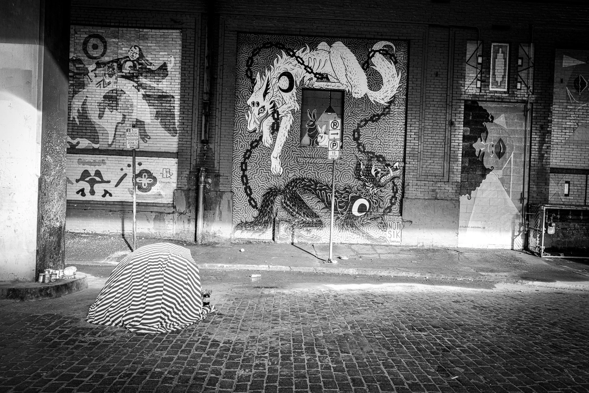 Street scene with graffiti murals on brick walls, including a dragon and human figures, a person sitting on the ground with a striped umbrella, and street signs in black and white.