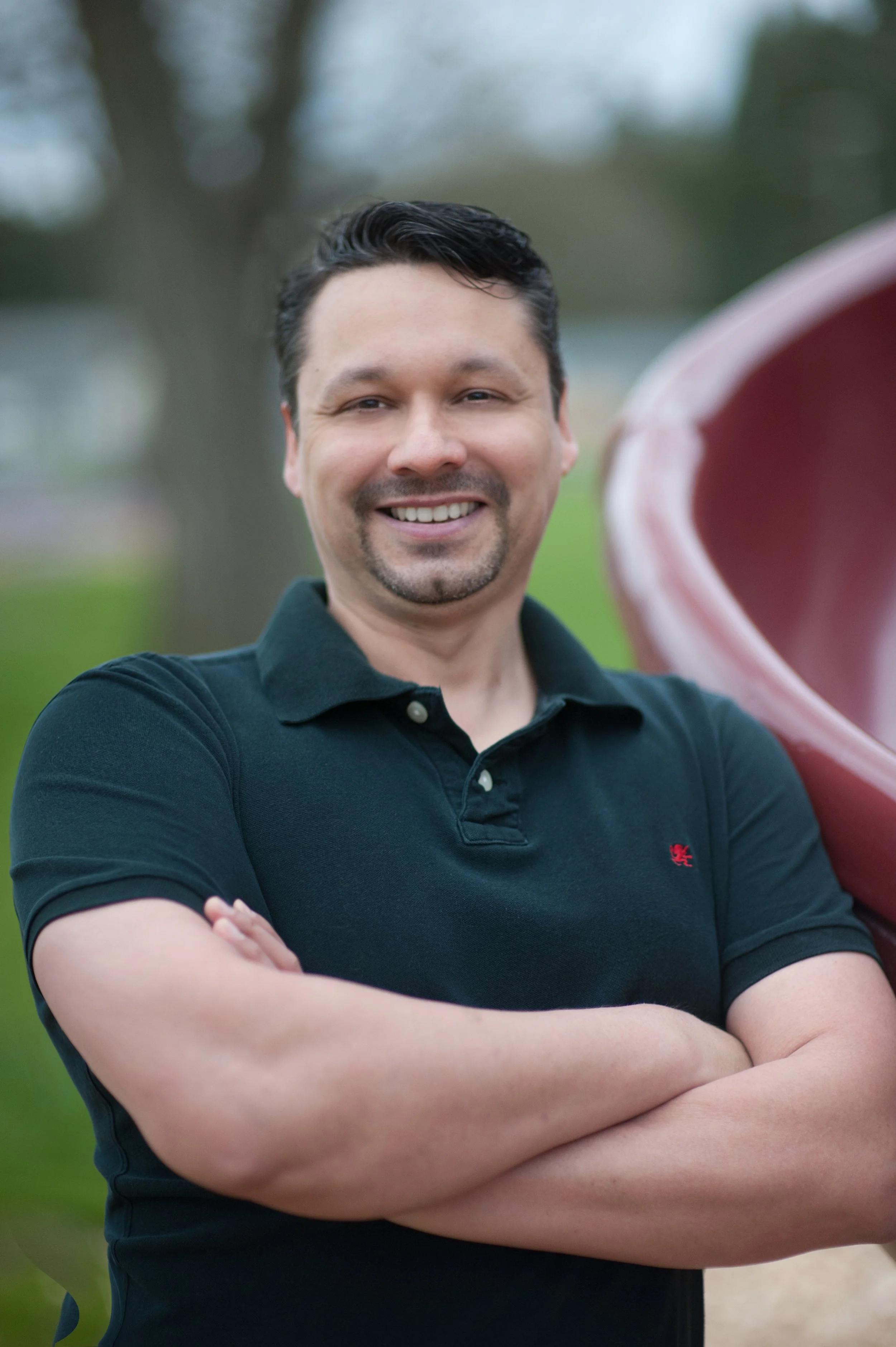 A man with short dark hair and a goatee smiling, wearing a black polo shirt with arms crossed, standing outdoors with blurred trees and grass in the background.