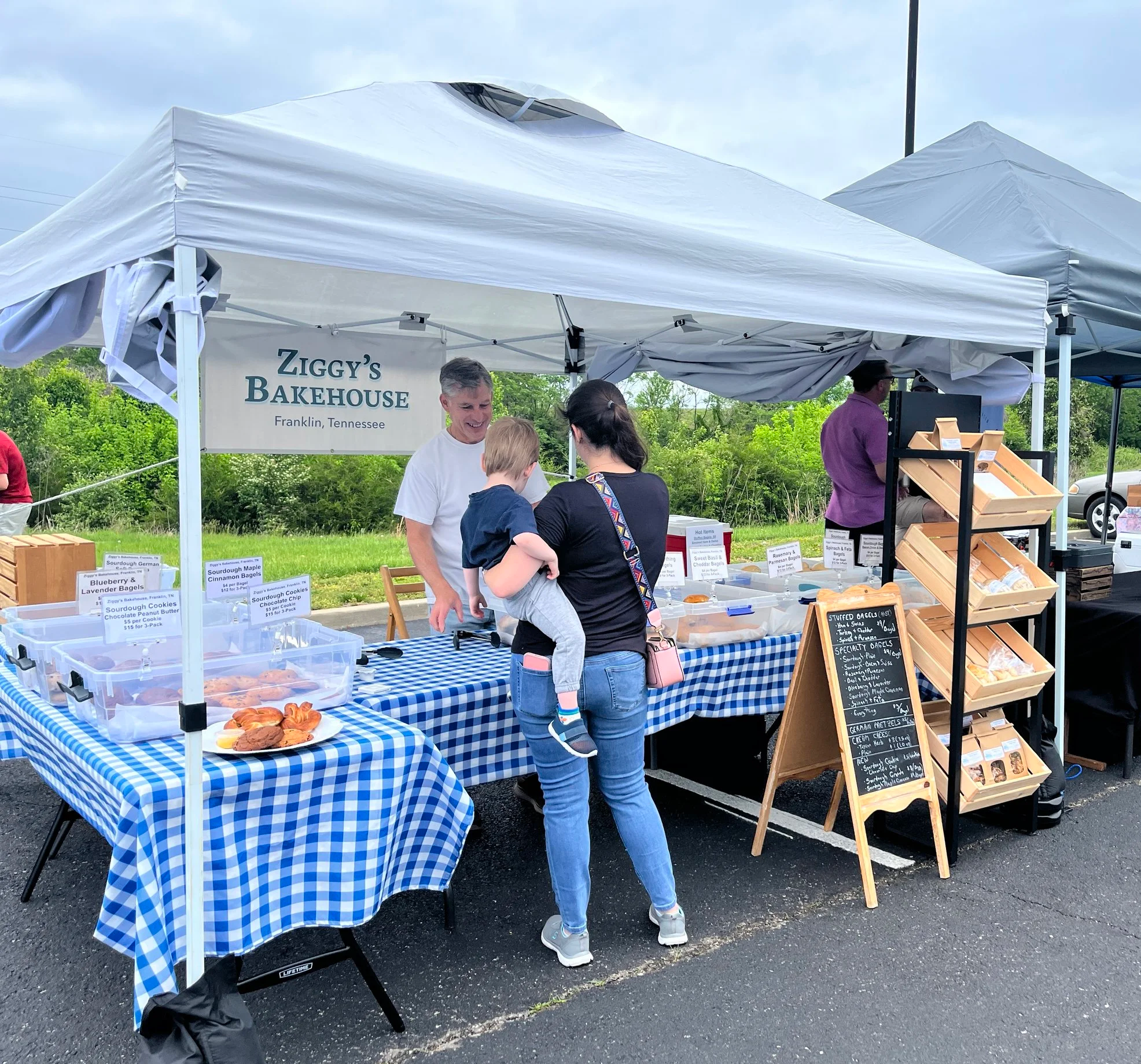People browsing baked goods at an outdoor market stall under a white canopy with a sign reading "Ziggy's Bakehouse" in Franklin, Tennessee. The stall displays cookies, bagels, and other baked items on blue and white checkered tablecloths.