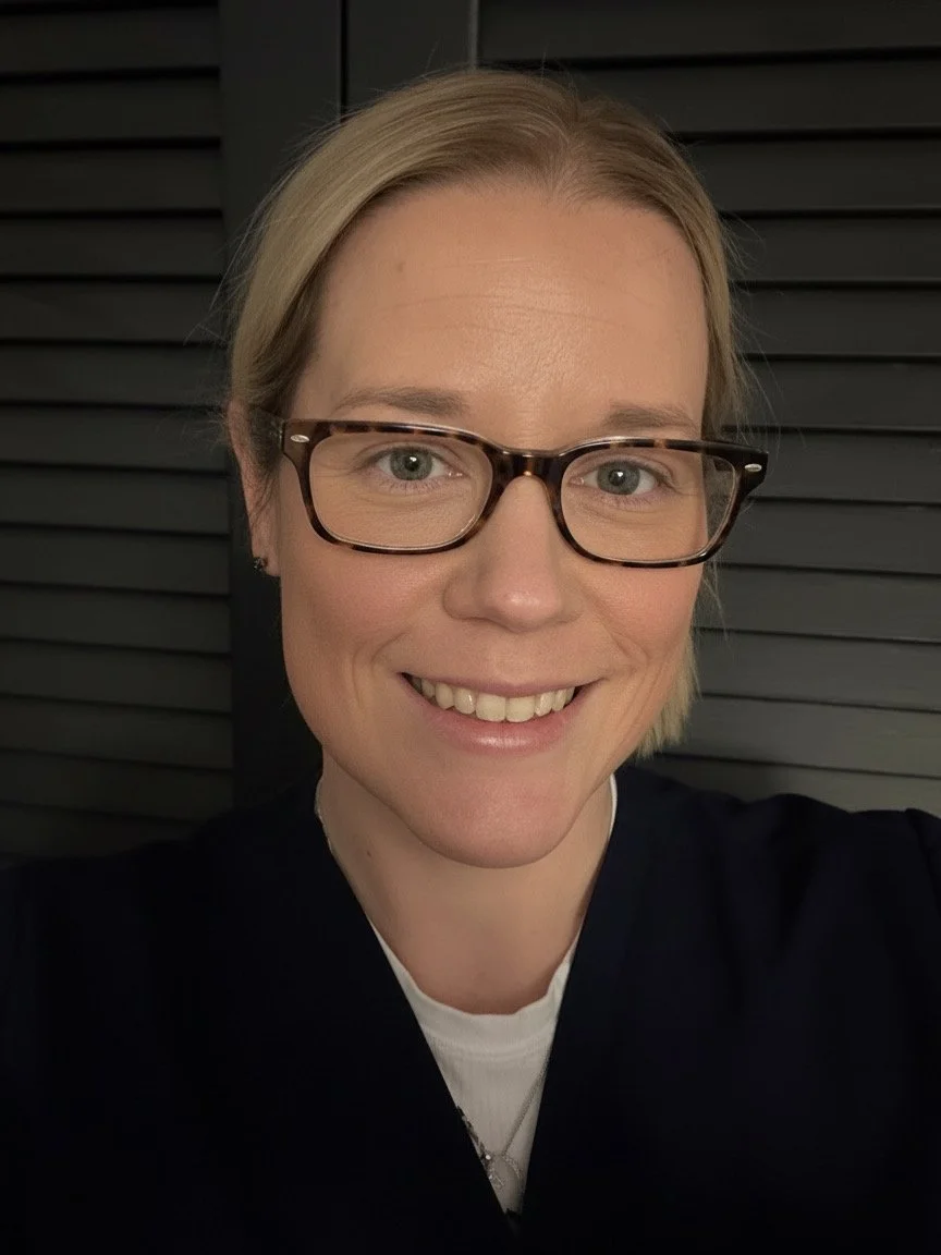 A woman with blonde hair, glasses, and a black top is smiling in front of a dark, slatted background. Belleame Aesthetics in Norwood, MA offers Botox, lip fillers, and dermal fillers with personalized treatments for natural-looking results.