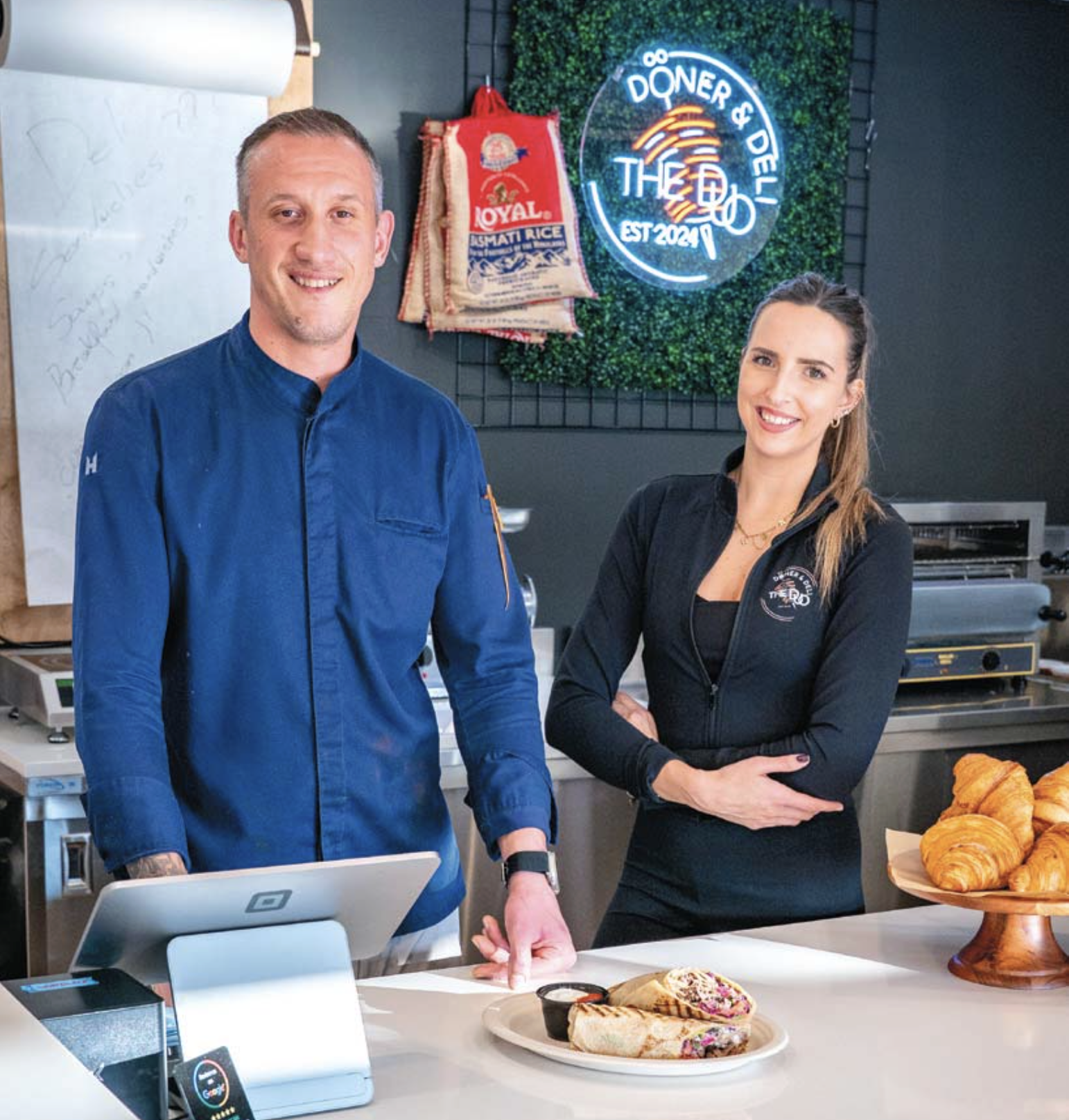 Two smiling restaurant staff members, a man and a woman, standing behind a counter with pastries, in a restaurant with a neon sign in the background that reads 'DÖNER & THEO' and a bag of rice hanging on the wall.