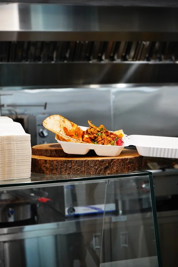 Close-up of a takeout container of food with chips, placed on a round wooden slice. There are paper napkins stacked beside it, on a countertop in what appears to be a kitchen or food service area.