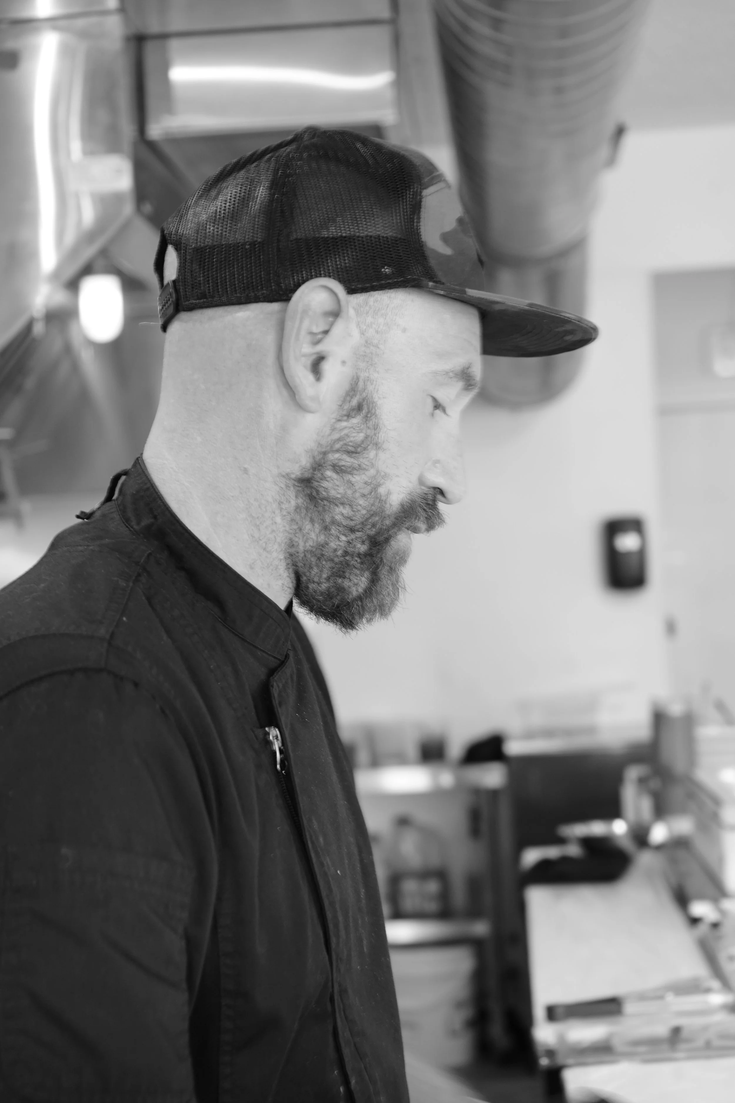 Side profile of a man with a beard wearing a black cap and black jacket, standing indoors with metal shelves and kitchen supplies in the background.