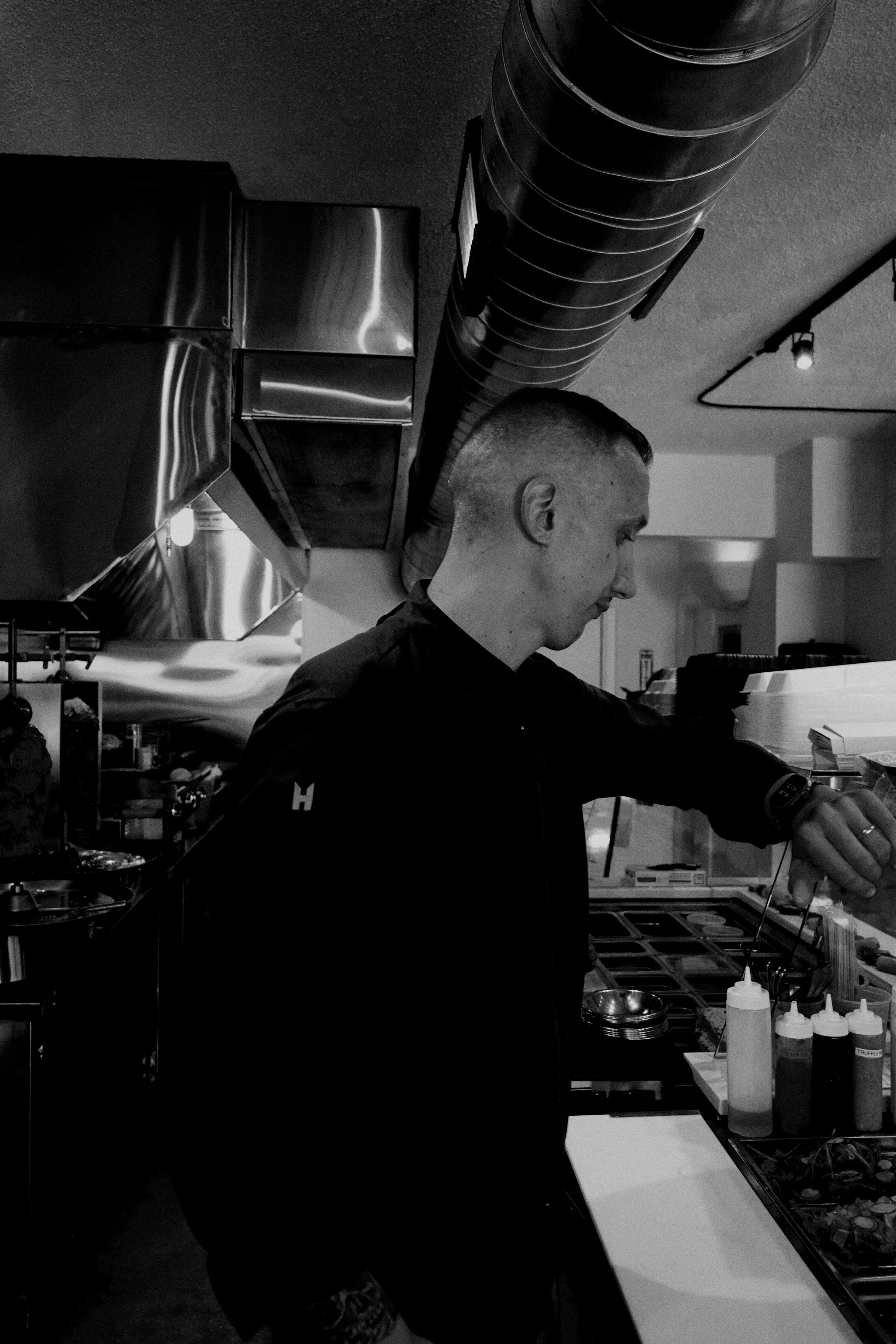 A chef in a black uniform is preparing food in a restaurant kitchen. The image shows various cooking utensils, condiment bottles, a stove, and a stainless steel ventilation hood. The chef is focused on cooking or plating dishes.