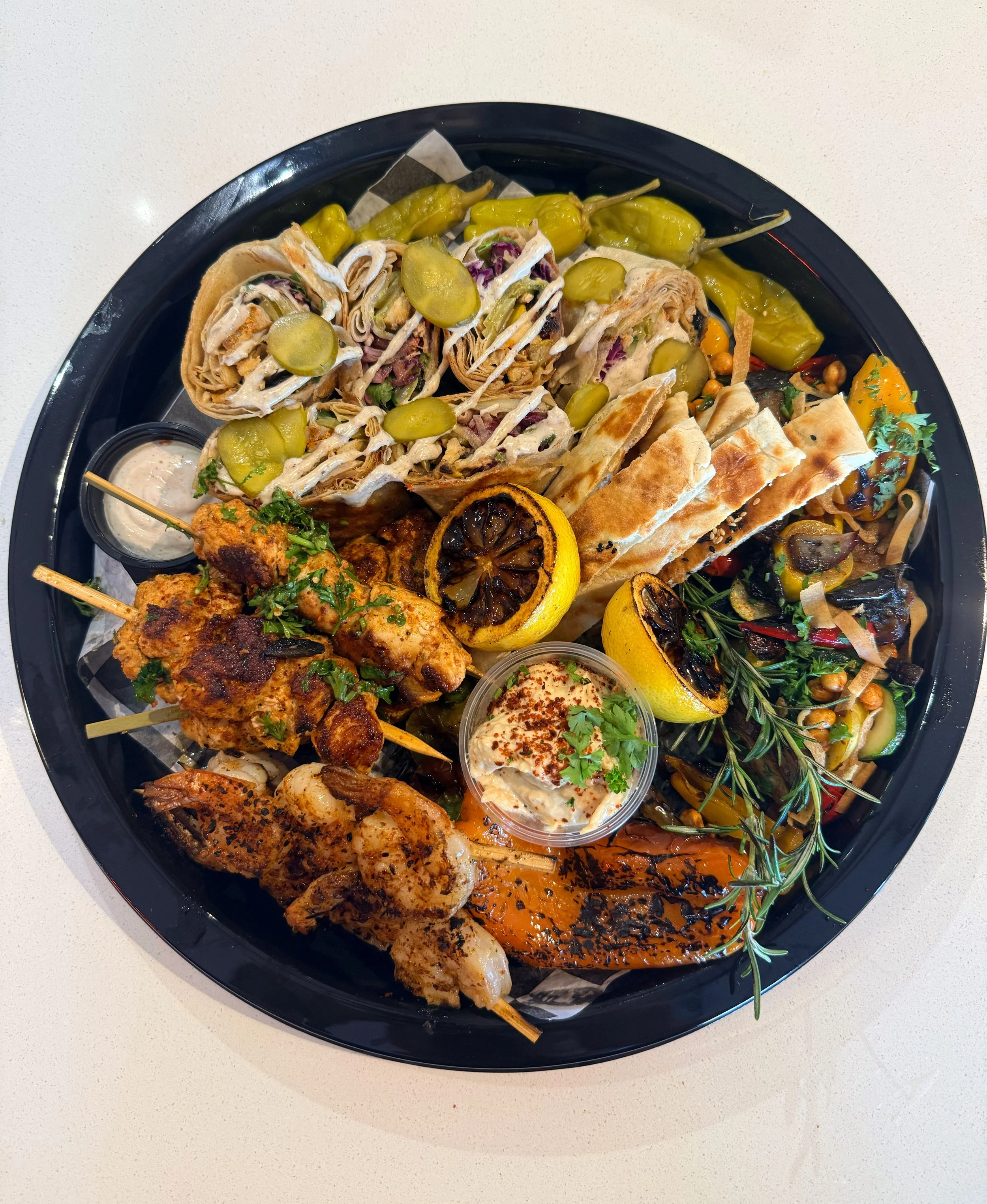 A black oval platter filled with various grilled meats, vegetables, wraps, and garnishes, including grilled lemon halves and fresh herbs.