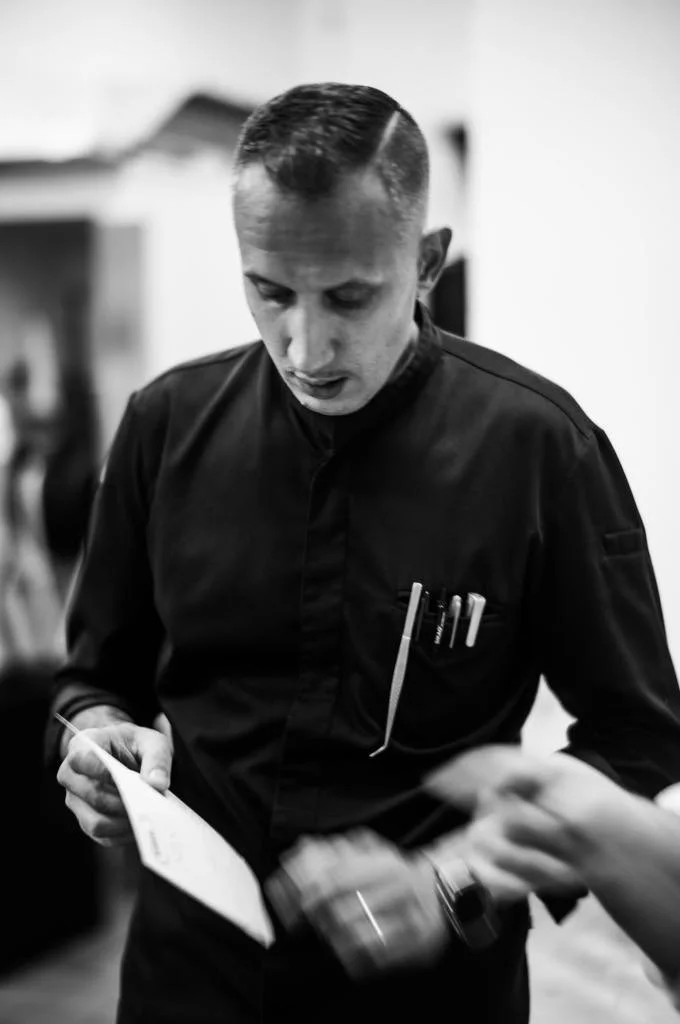 A male chef in a black uniform looking down and working in a kitchen.