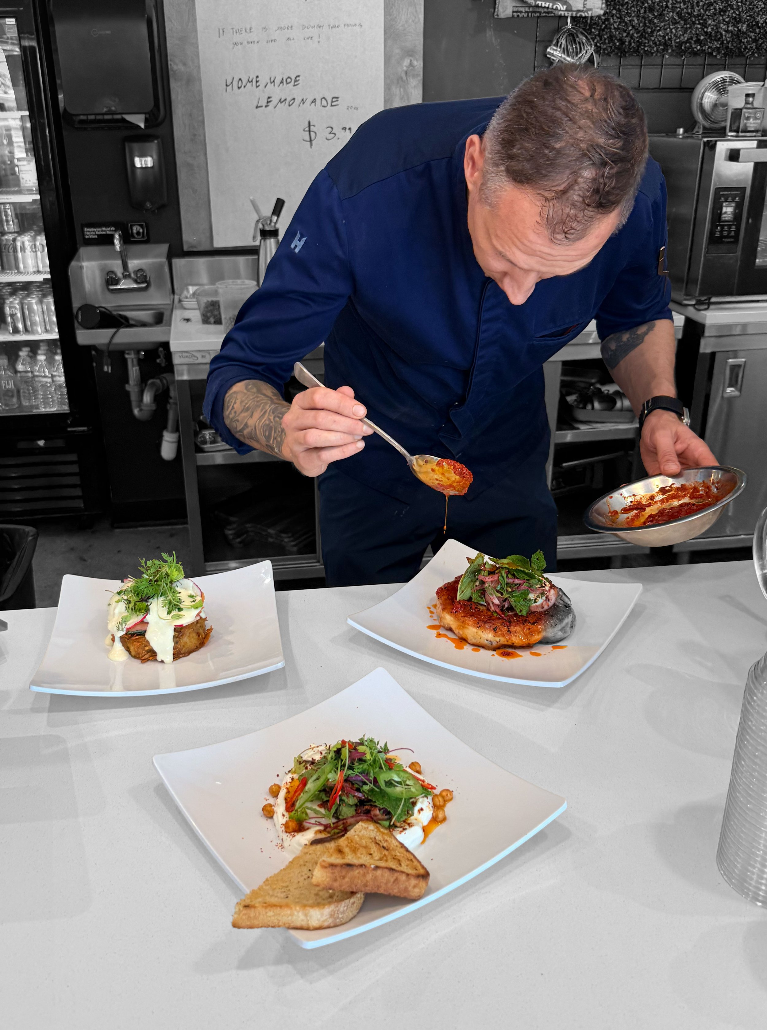 A man with tattoos on his arm wearing a blue chef's jacket is preparing food in a kitchen. On the table in front of him are three white square plates with various dishes, including salads, grilled fish, and toasted bread.