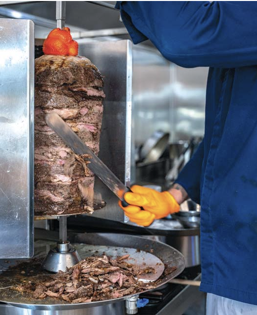 A person wearing a blue chef's coat and orange gloves slices a large rotisserie roast beef with a knife, with the roast beef cooked on a vertical spit and some slices on a plate below.