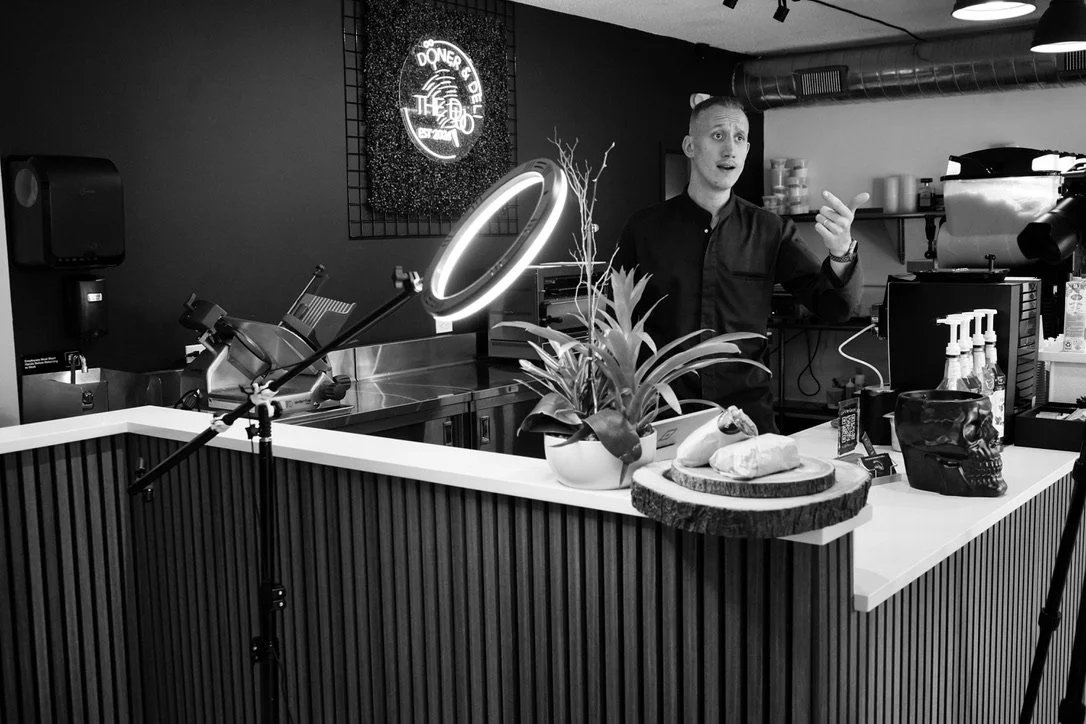 A man is speaking behind a counter with a potted plant, skull sculpture, and food items on it in a modern restaurant or cafe. There is a ring light and photography equipment around.