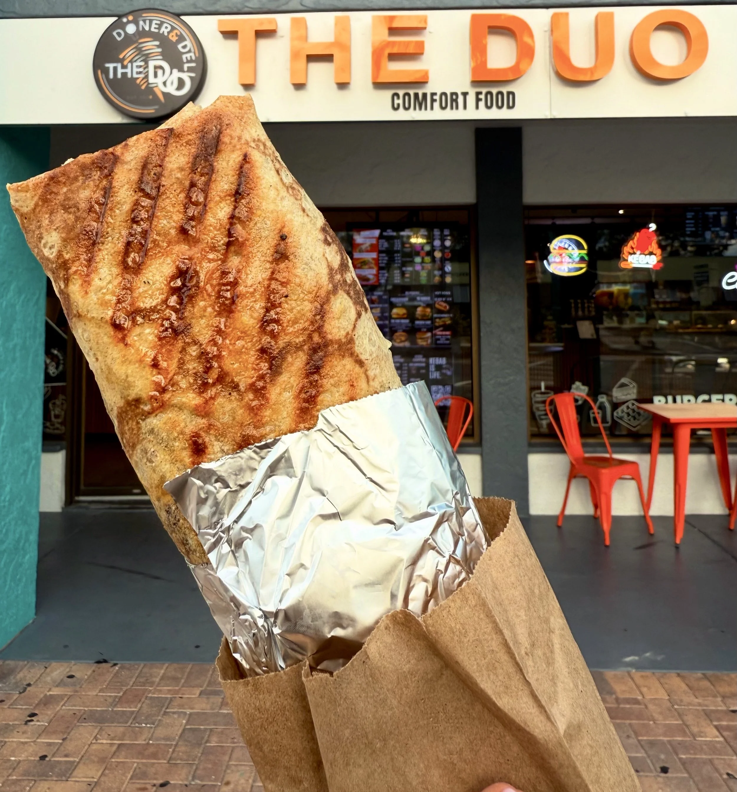 Close-up of a grilled burrito wrapped in aluminum foil and resting on a paper bag, in front of a fast food restaurant with a sign that reads 'The DUO Comfort Food.'