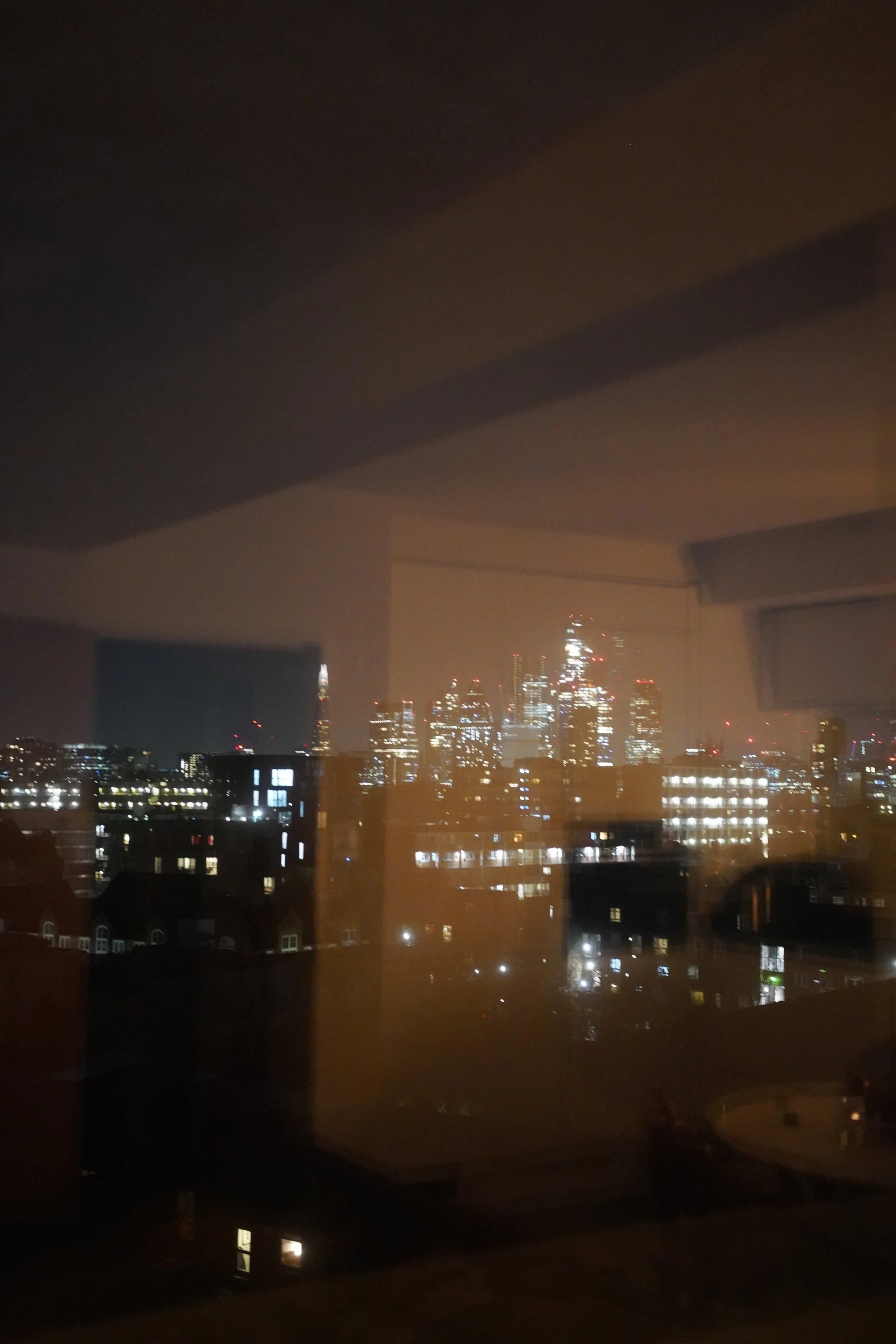 Night view of a city skyline through a window, with illuminated buildings and a dark sky, reflected in the glass.