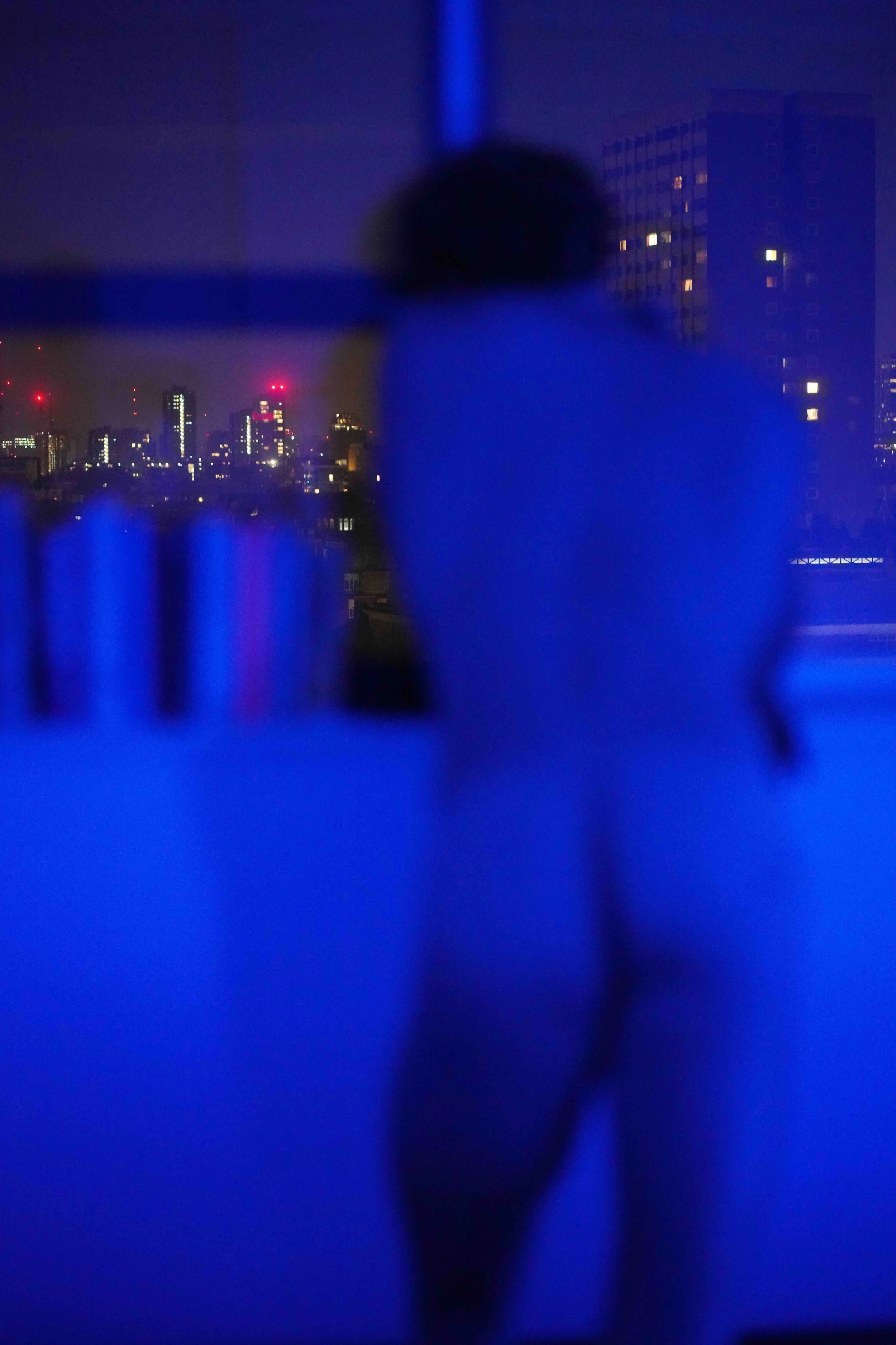 A blurry night cityscape with tall buildings and illuminated windows, viewed through a glass or window with blue lighting and reflections.