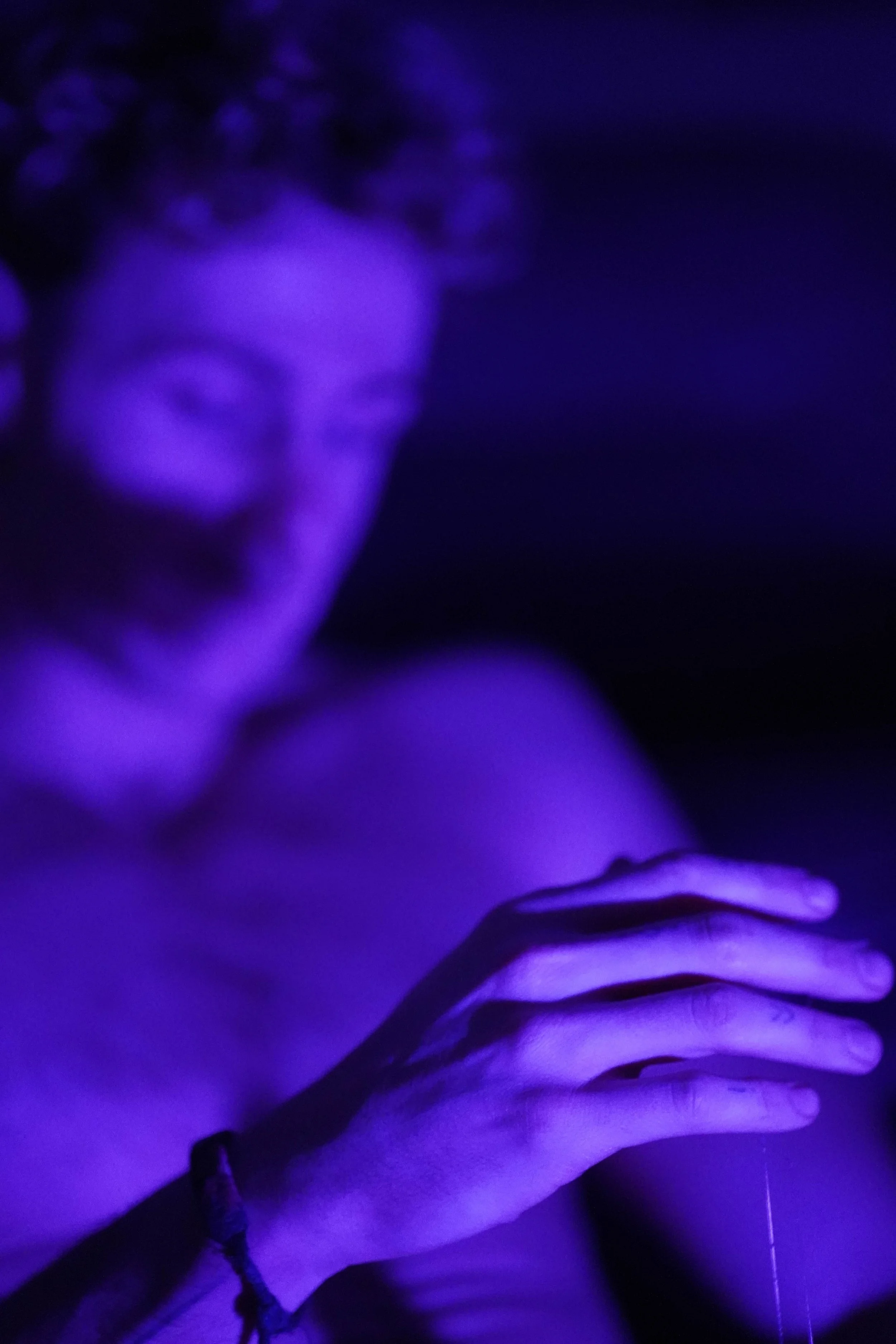 A person with pink hair, wearing glasses, depicted in purple lighting, touching their chest with their hand.