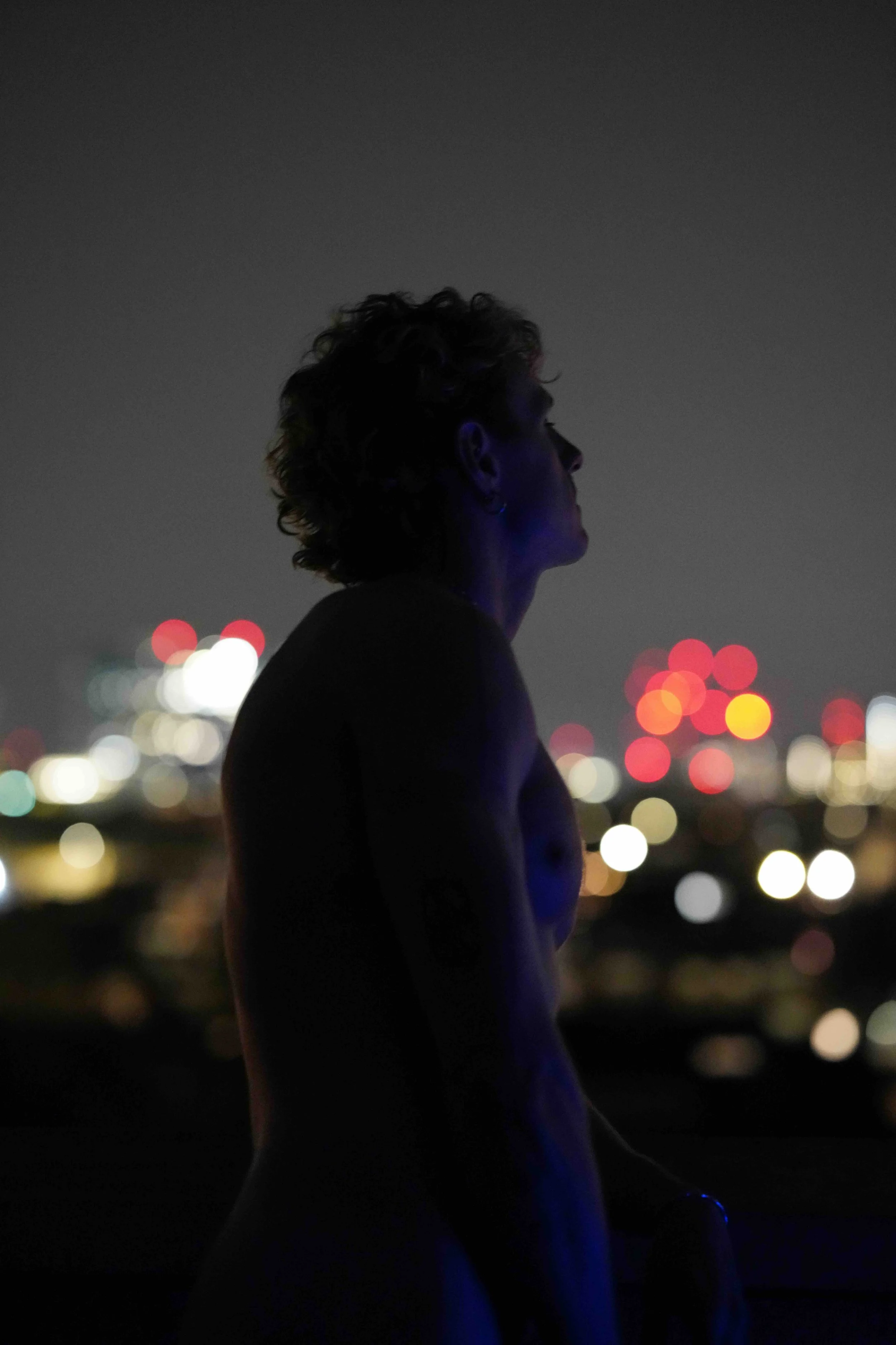 Silhouette of a woman with short curly hair looking to the right, against a blurred cityscape at night with colorful bokeh lights.