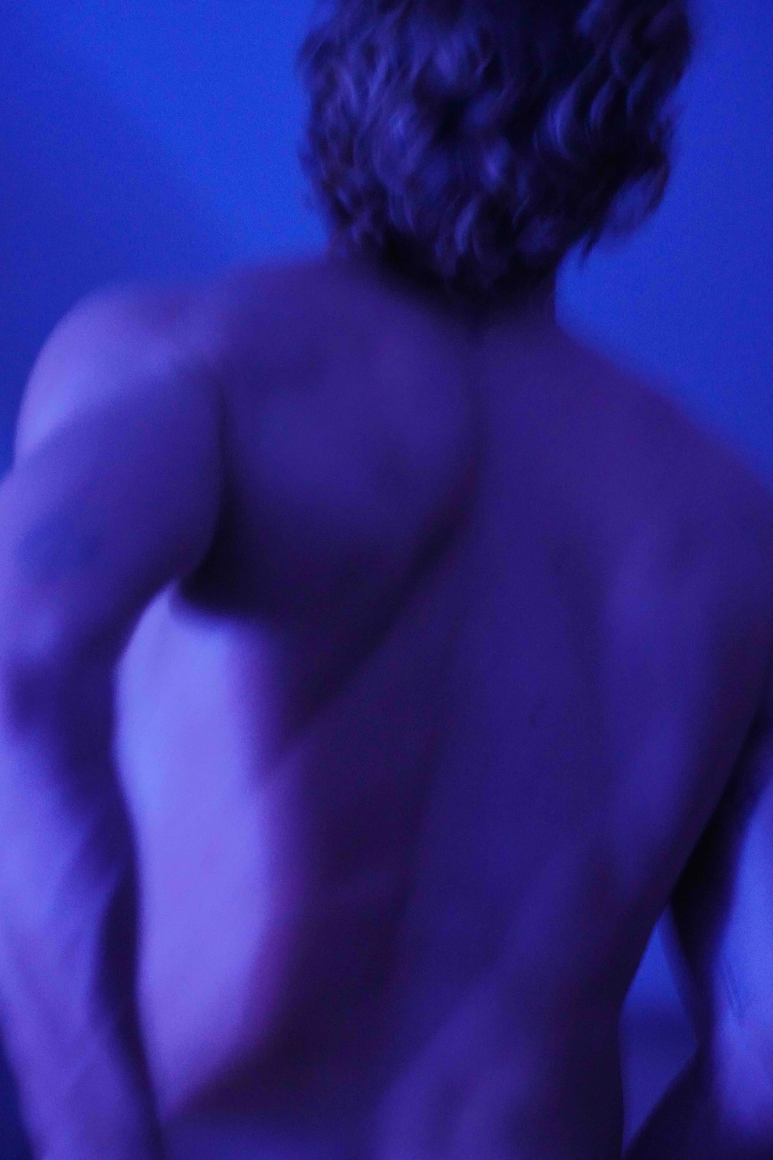 Close-up of a person's shoulder and back, with curly hair, illuminated in blue lighting.