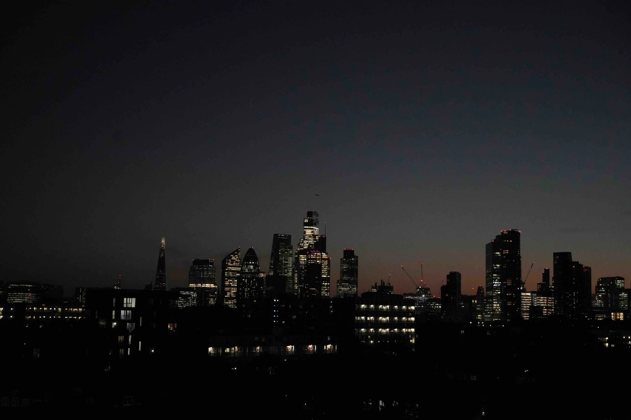 Nighttime city skyline featuring tall skyscrapers with illuminated windows and construction cranes against a dark sky.