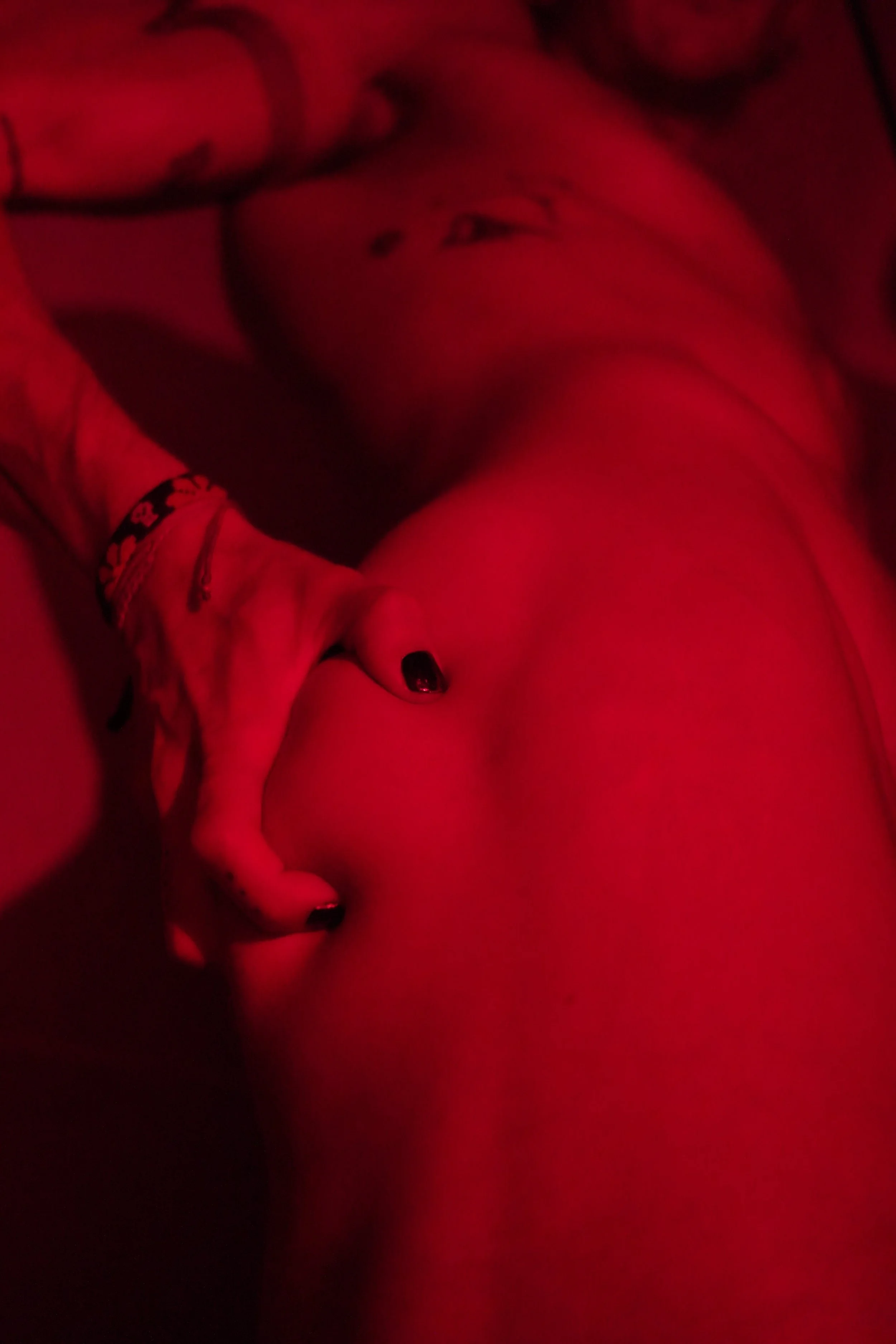 A person lying on their back with their hand on their lower stomach, wearing a wristband and black-painted nails, illuminated by red lighting.
