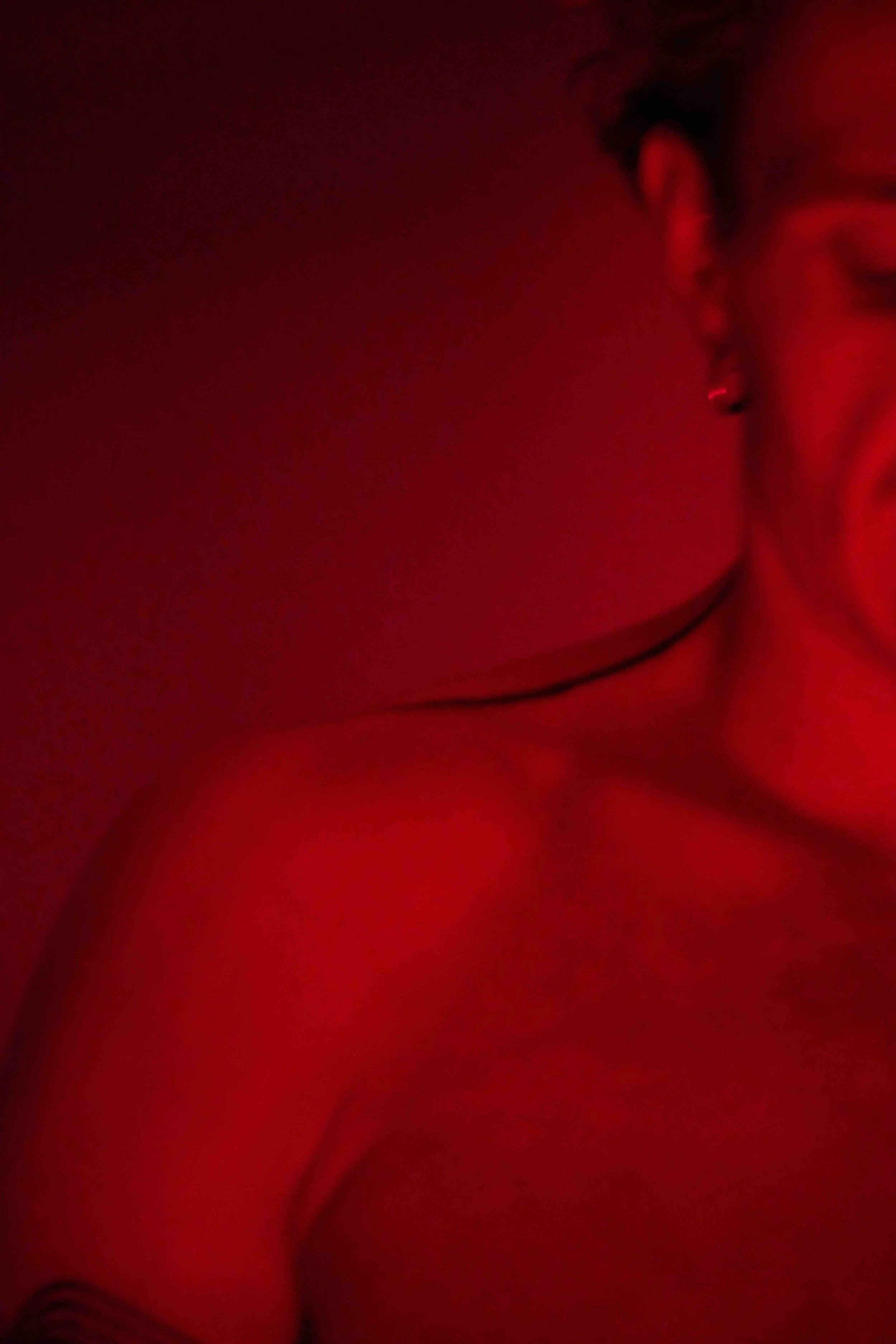 Close-up shot of a person's face and neck in red lighting with visible earrings, earrings, and a thin black necklace.