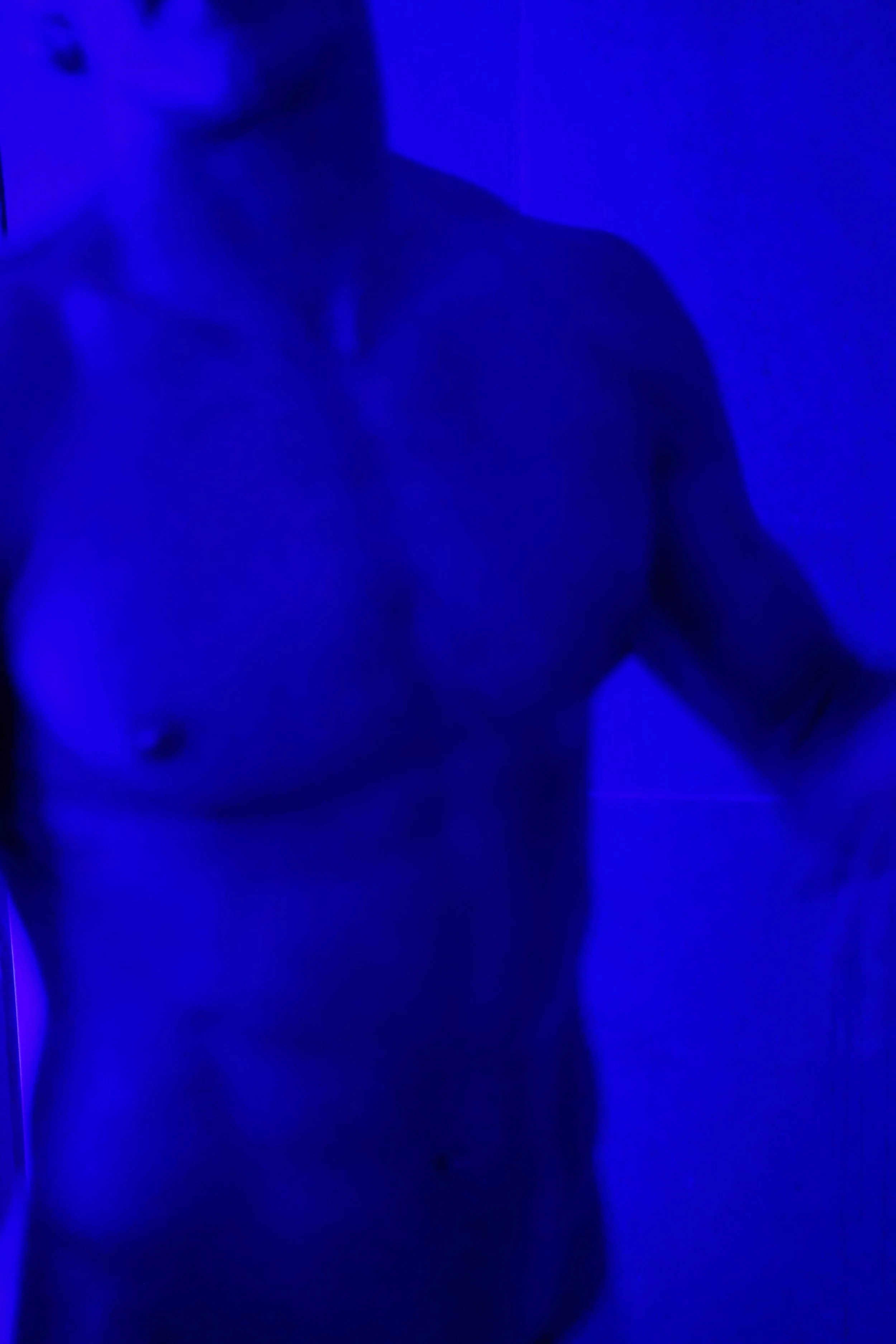 A shirtless man with visible chest and shoulder muscles, illuminated by blue light.