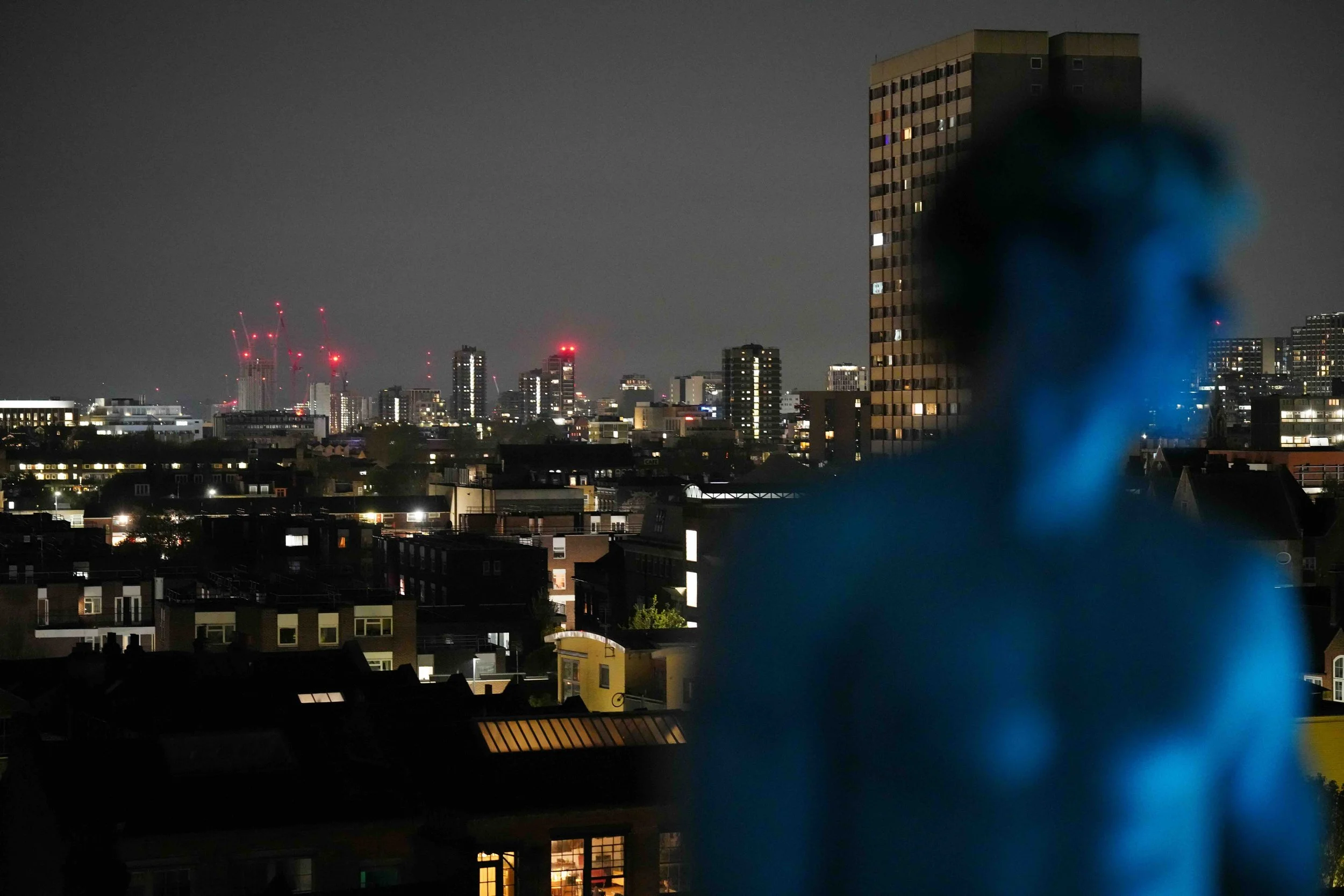 Nighttime cityscape with illuminated buildings, construction cranes, and a blurred figure in the foreground.