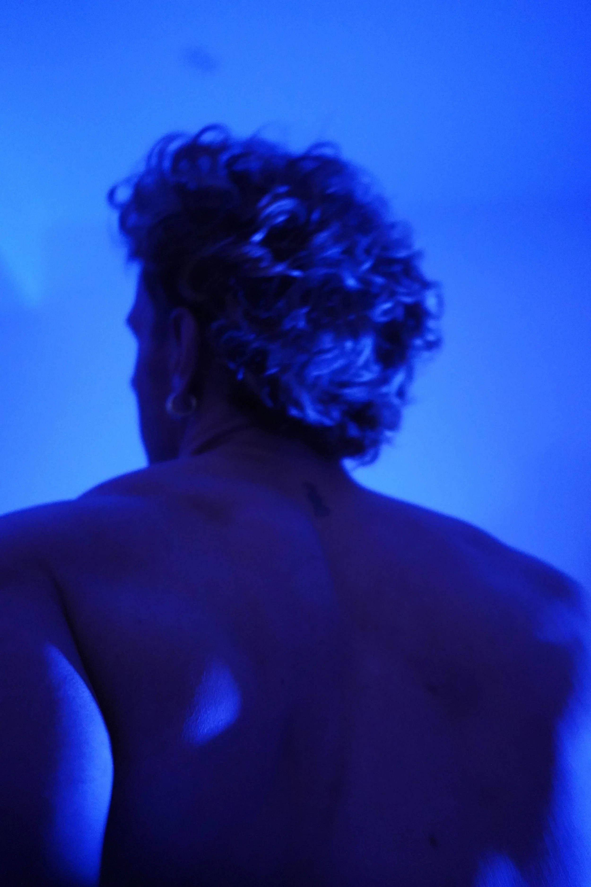 Back of a person's head with curly gray hair illuminated by blue light, showing their shoulder and upper back, against a blue background.