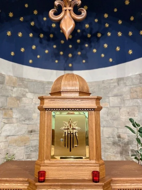 A wooden tabernacle with a gold door featuring a cross, set on a wooden surface with two red candles in front. The background includes a stone wall and a blue ceiling decorated with gold stars and a decorative gold ornament hanging above.