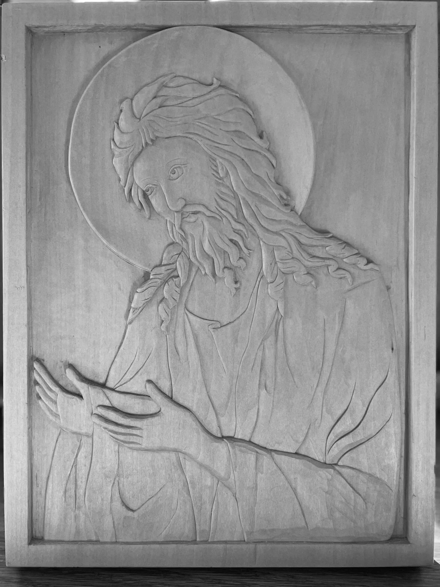 Wooden carved relief of Jesus Christ with a halo, long hair, and beard, gesturing with hands near his chest, in a religious style.