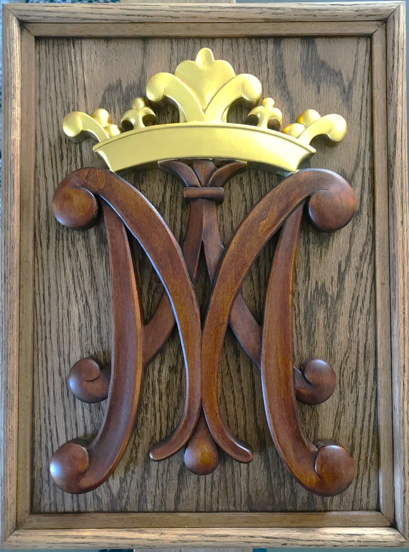 Wooden plaque featuring a carved monogram of the letter 'W' topped with a golden crown, mounted on a wooden background.