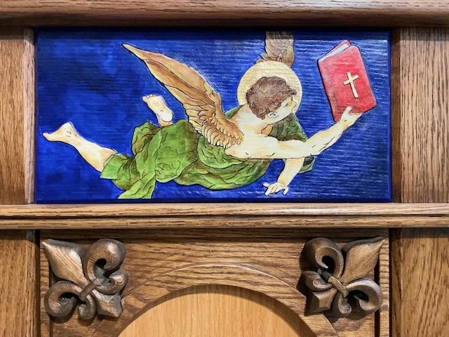 Relief artwork of a flying angel with wings, holding a red book with a cross, on a blue background, mounted on a wooden surface.