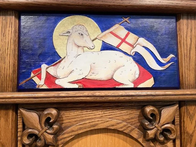 Religious wood carving depicting a lamb with a halo, lying on a red cloth, holding a flag with a red cross, against a dark blue background.