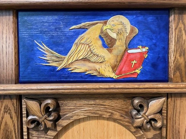 A carved wooden panel with a painted illustration of an eagle holding a Bible with a cross, set against a blue background.